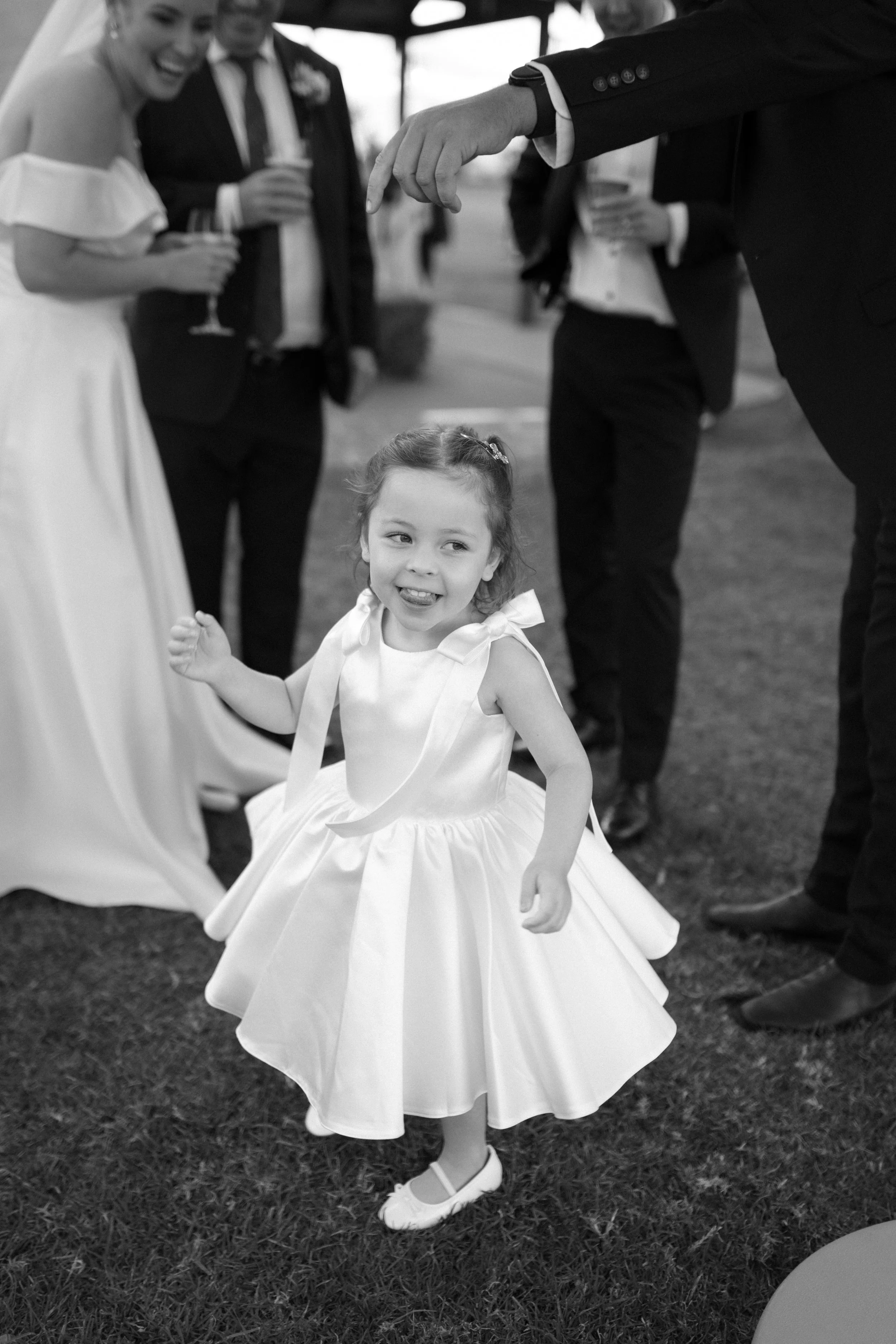 A young girl in a fancy dress and shoes dancing or posing at a wedding or formal event, surrounded by adults in suits holding drinks, with a blurred outdoor background.