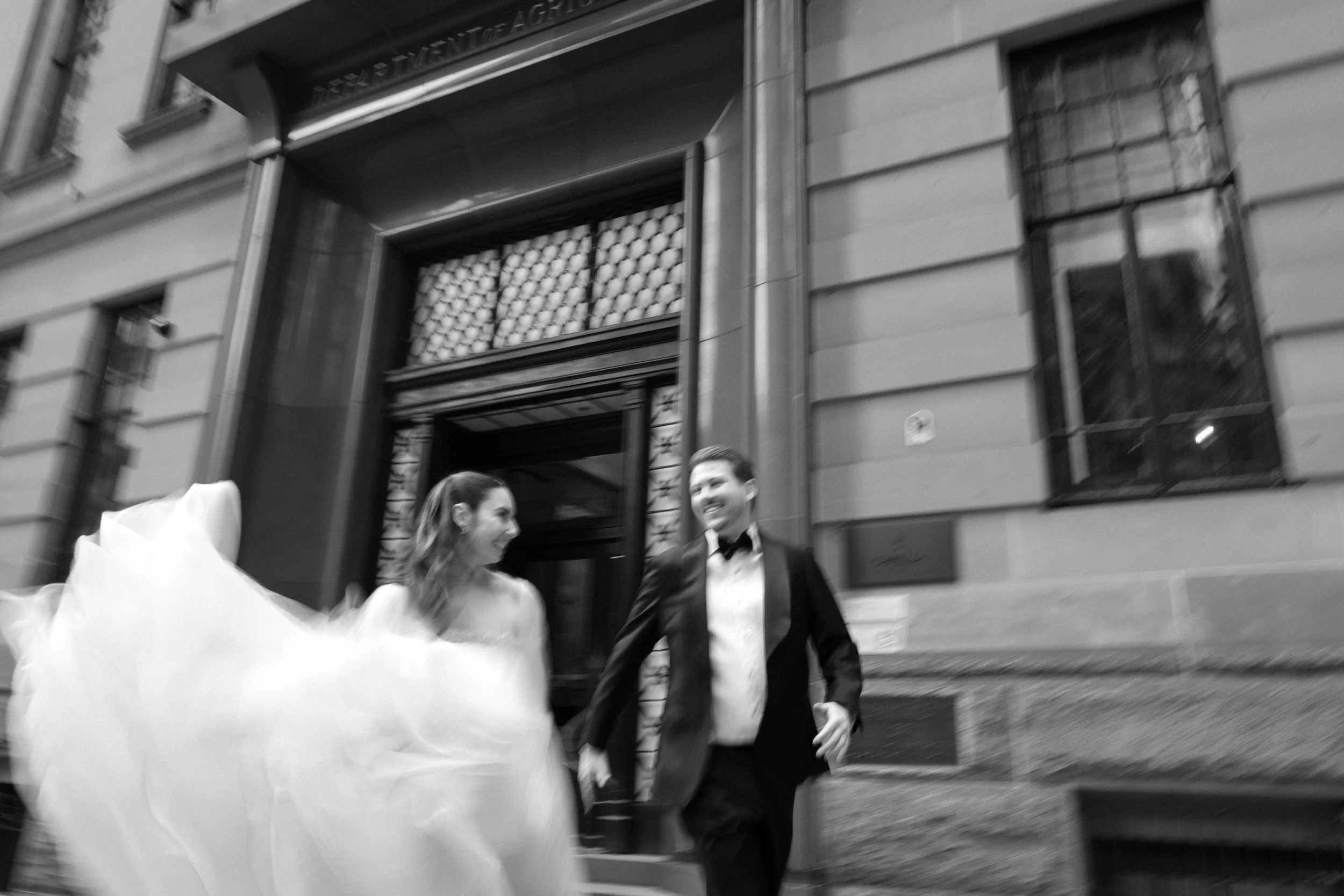A bride and groom in wedding attire smiling and walking outside a building, with the bride's dress flowing.