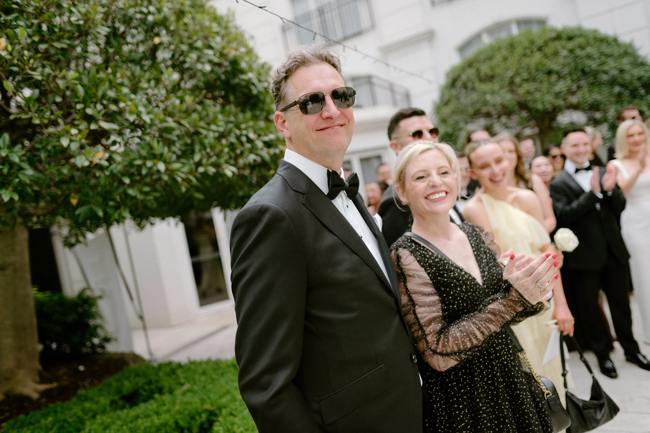 A group of people dressed in formal attire at an outdoor event, with one man in a tuxedo and sunglasses smiling in the foreground, and a woman wearing a black polka dot dress clapping beside him, surrounded by other elegantly dressed attendees.