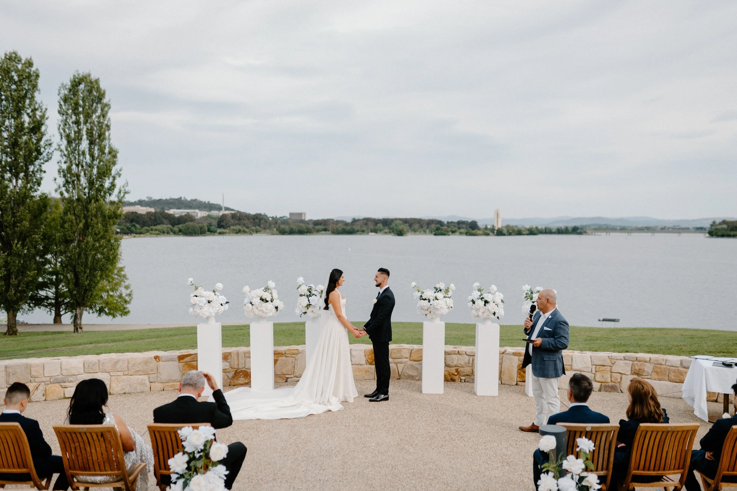 Wedding ceremony outdoors by a lake with a bride and groom holding hands, officiant speaking, and guests seated on chairs facing the couple.