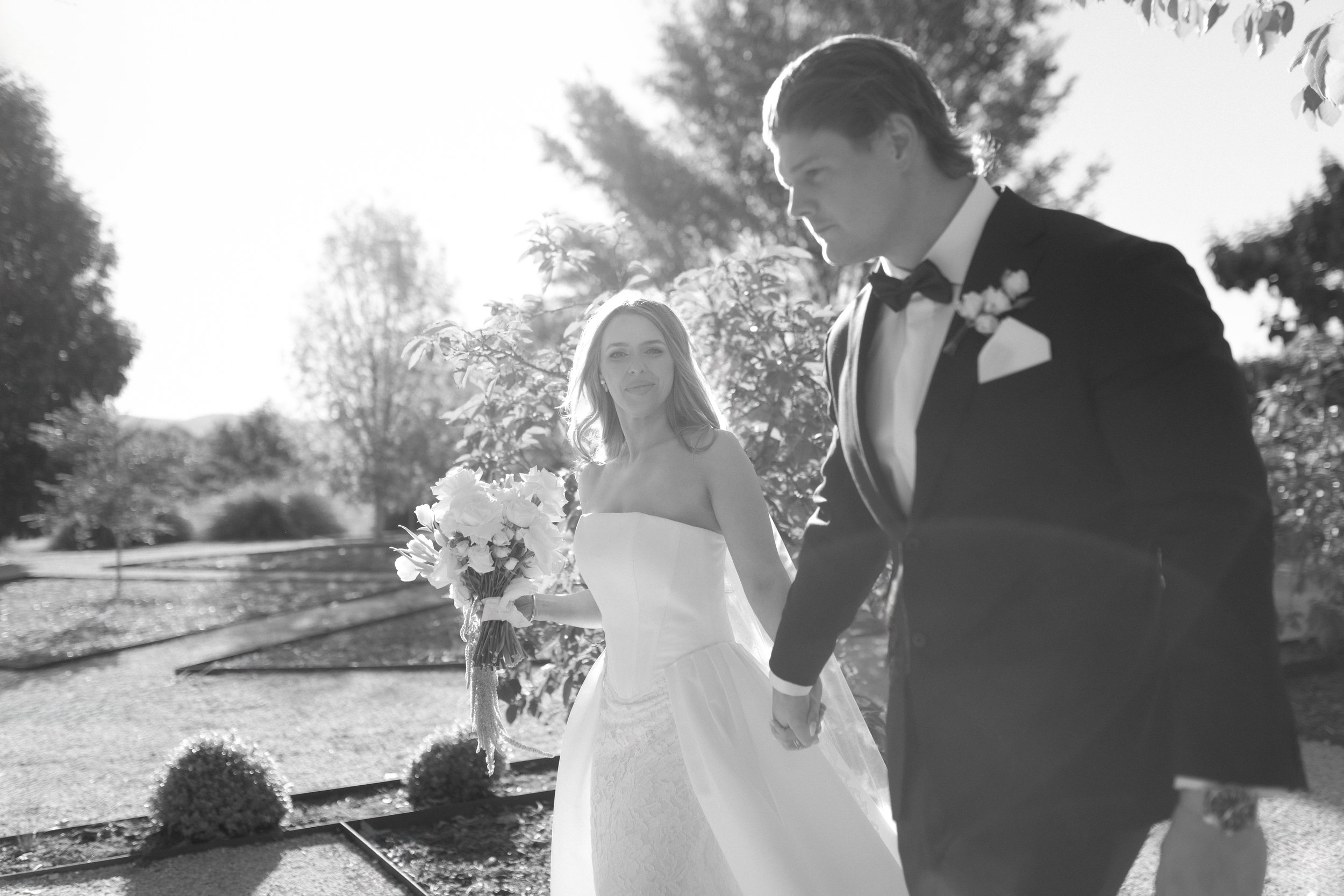 A black-and-white photo of a bride and groom holding hands outdoors. The bride is holding a bouquet of flowers and smiling softly, while the groom is looking at her with a serious expression. The background features trees and a bright sky.