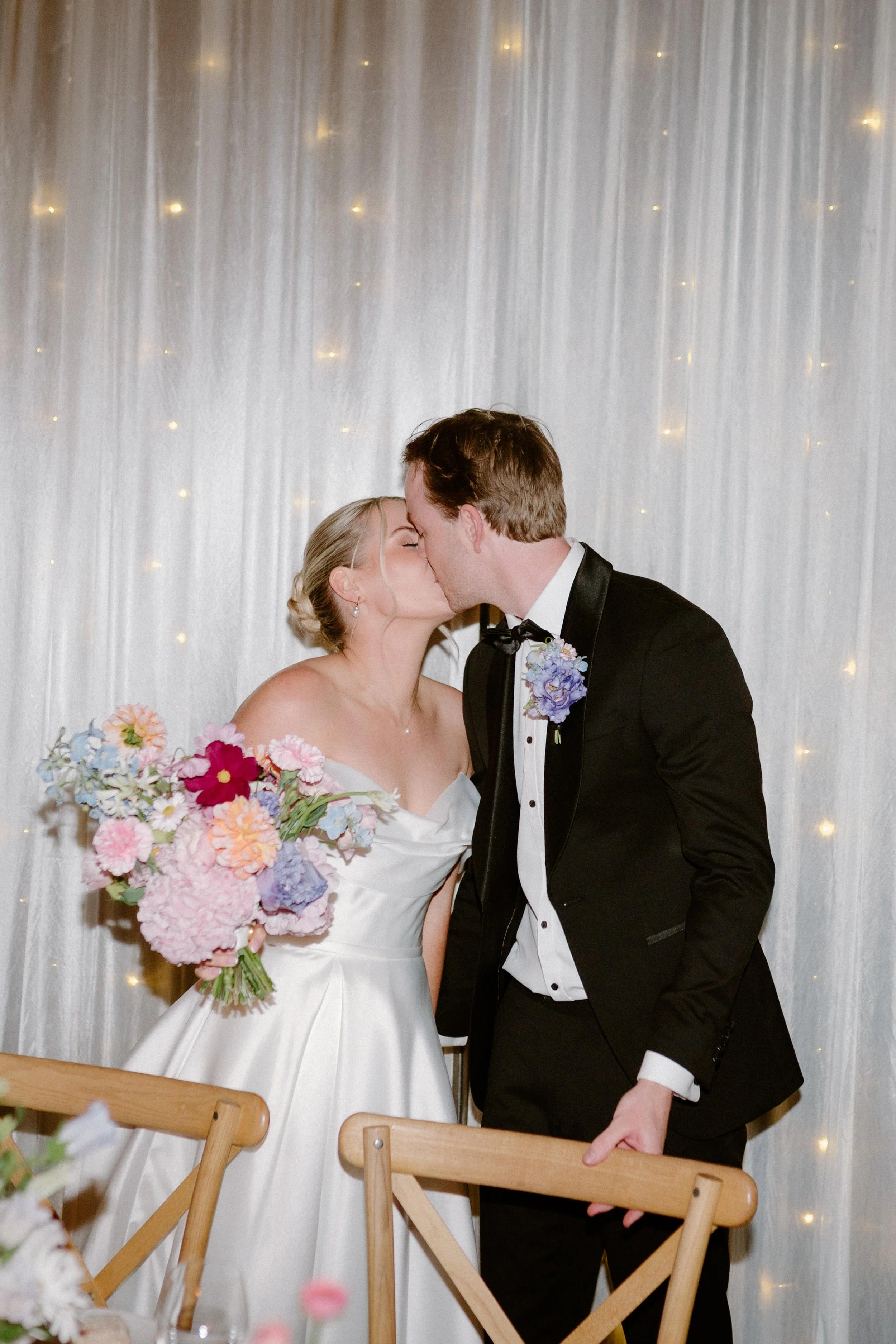 A bride and groom sharing a kiss at their wedding reception, standing in front of a white curtain with small fairy lights. The bride is holding a large bouquet of colorful flowers.