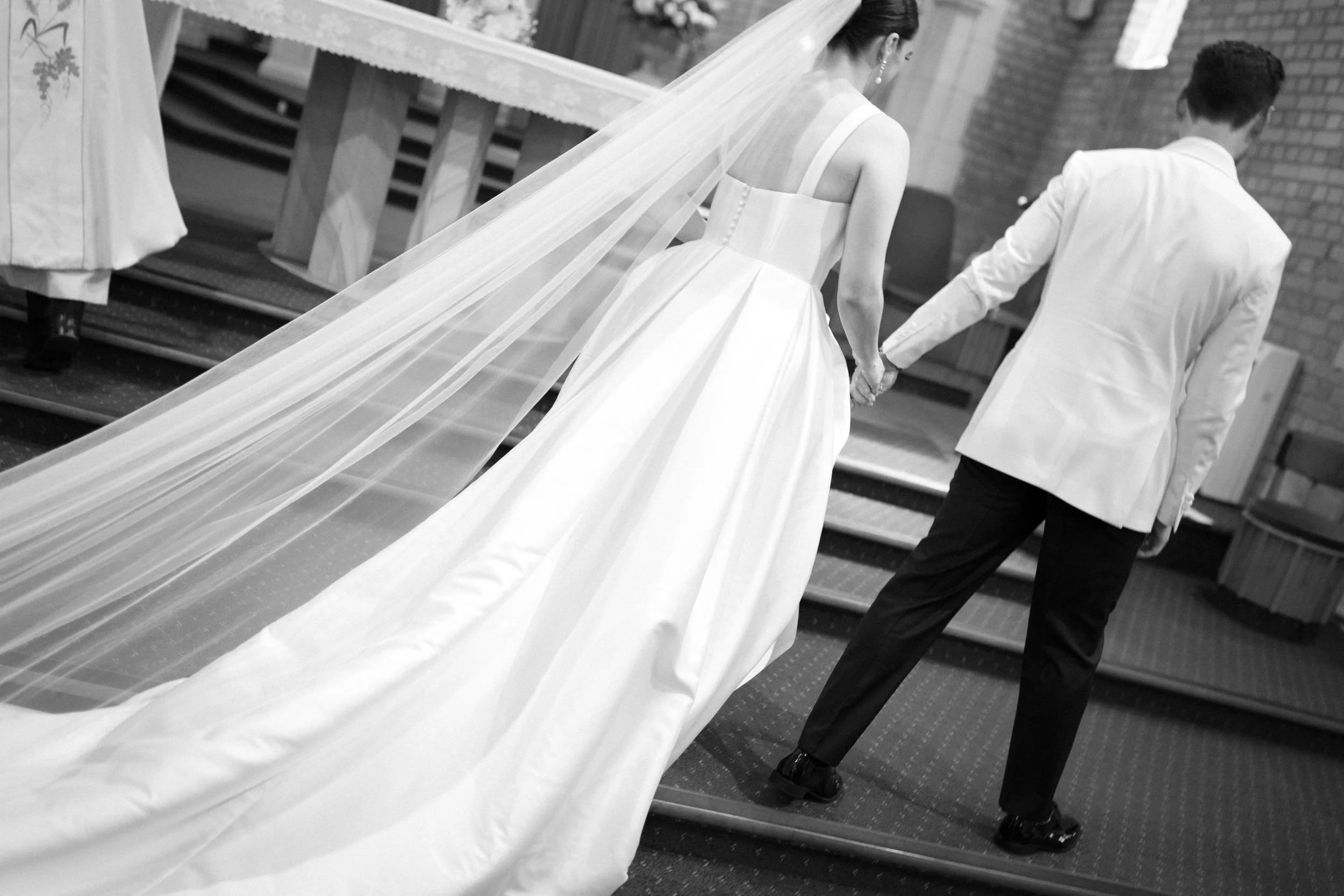 A bride and groom holding hands during their wedding ceremony in a church, with the bride wearing a long white wedding dress and veil and the groom in a white jacket and black pants, reflected in a mirror.