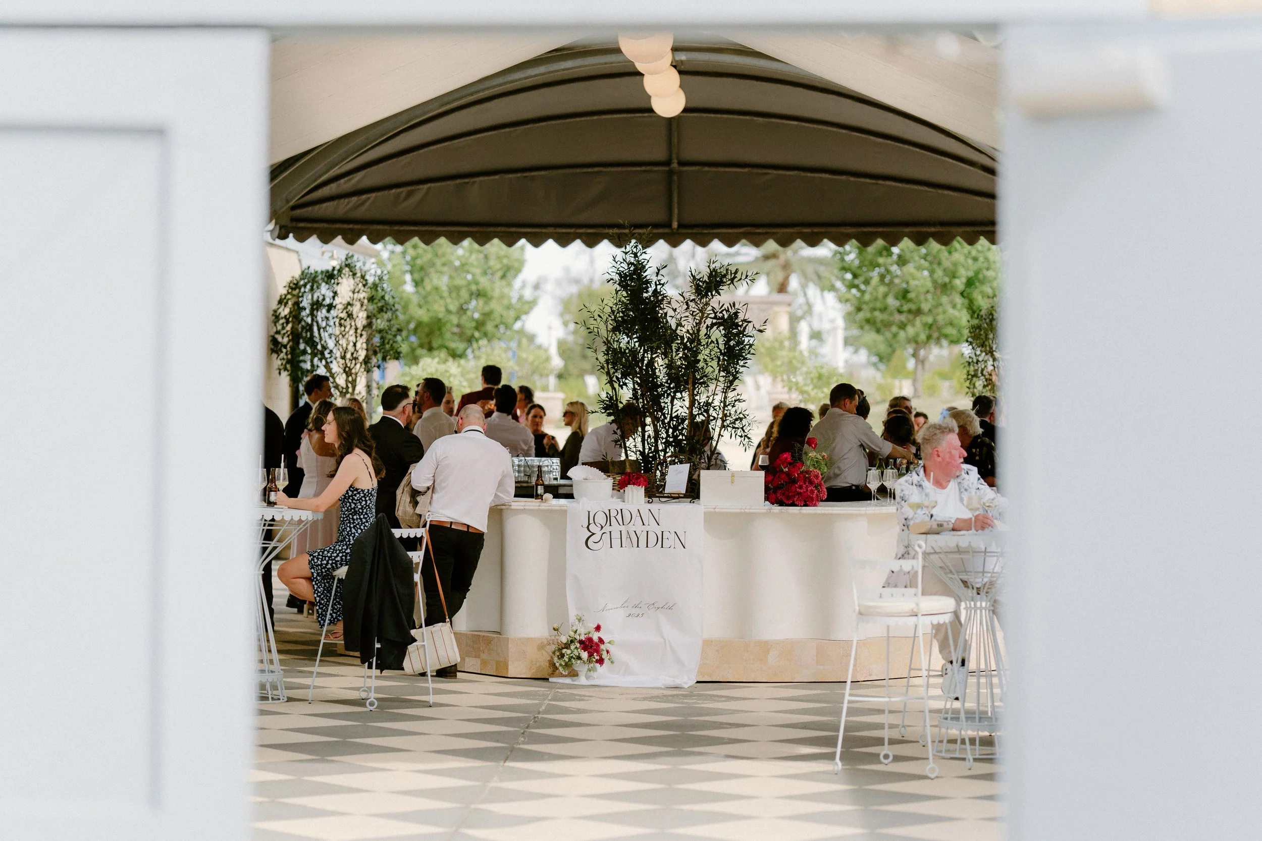 The Sienna, Hunter Valley Wedding Venue, Wedding Photos. People gathered at a wedding reception under a tent, with a sign reading 'Jordan & Hayden' on a white drape, floral arrangements, and a checkered tile floor.