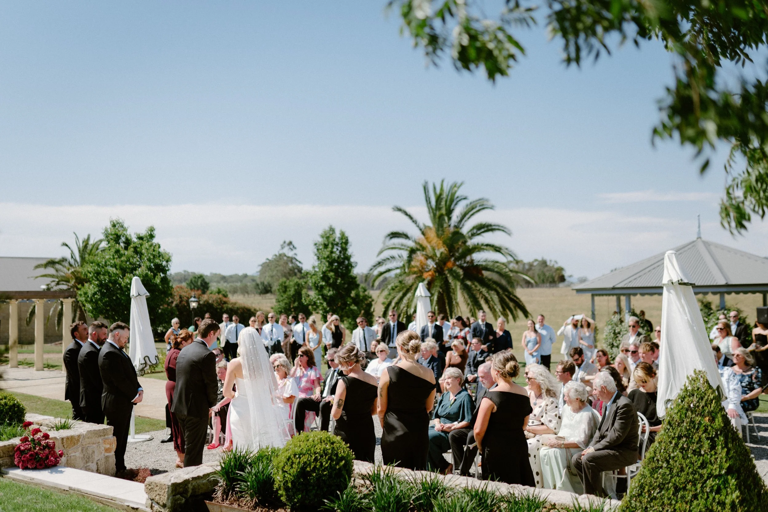 A wedding ceremony outdoors with a bride and groom standing in front of guests, under a clear sky with trees and a pavilion nearby.