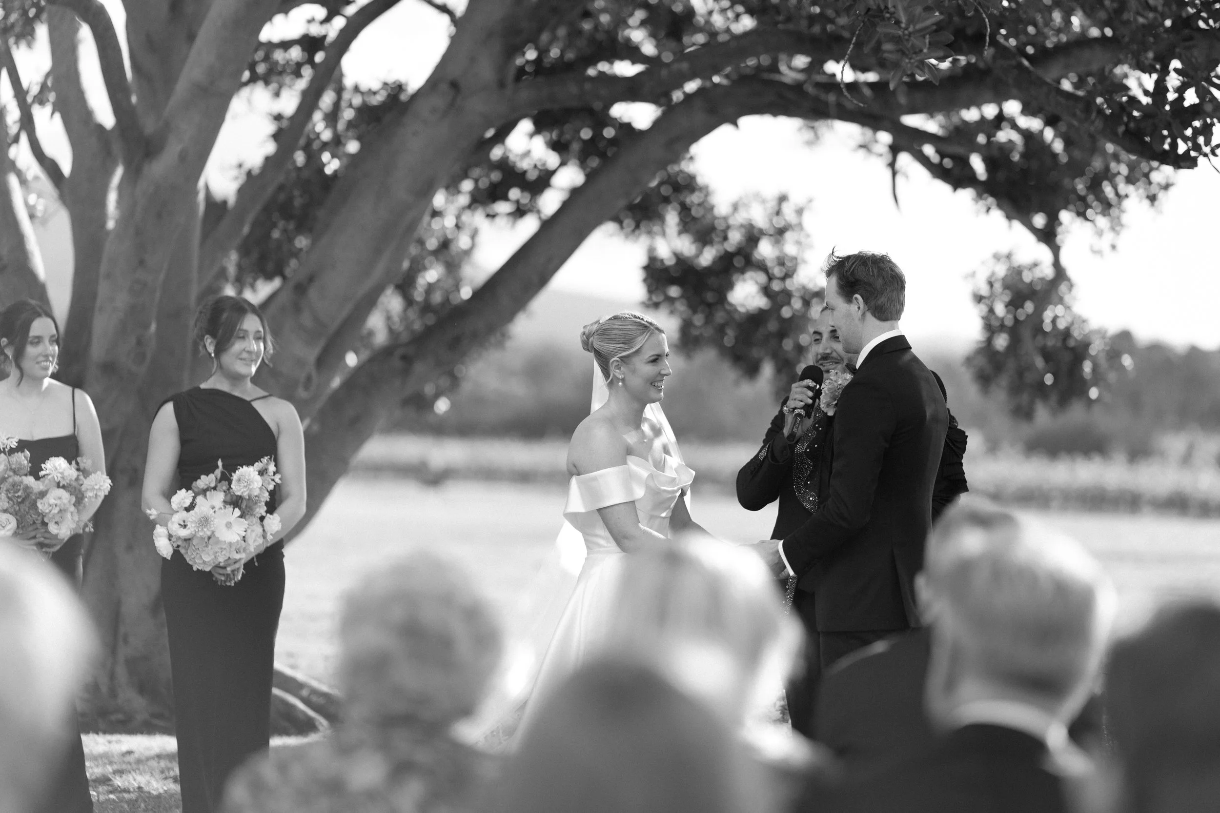 A black and white photo of a wedding ceremony outdoors, with a bride and groom exchanging vows, surrounded by bridesmaids holding bouquets, under large trees.