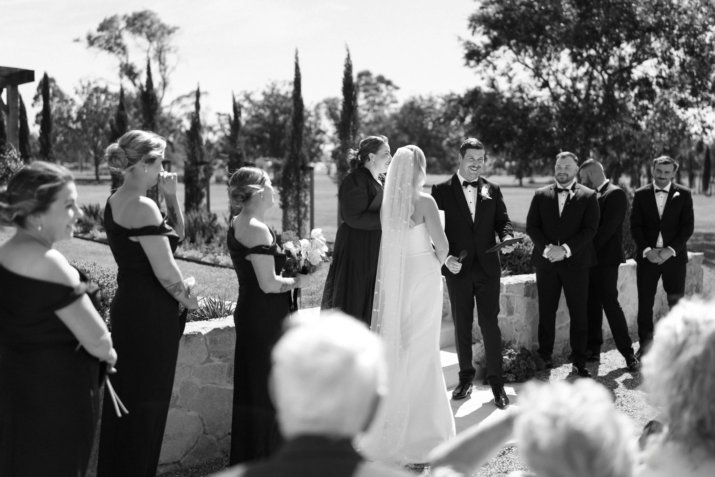Black and white photo of a wedding ceremony outdoors with the bride and groom surrounded by wedding party and guests.