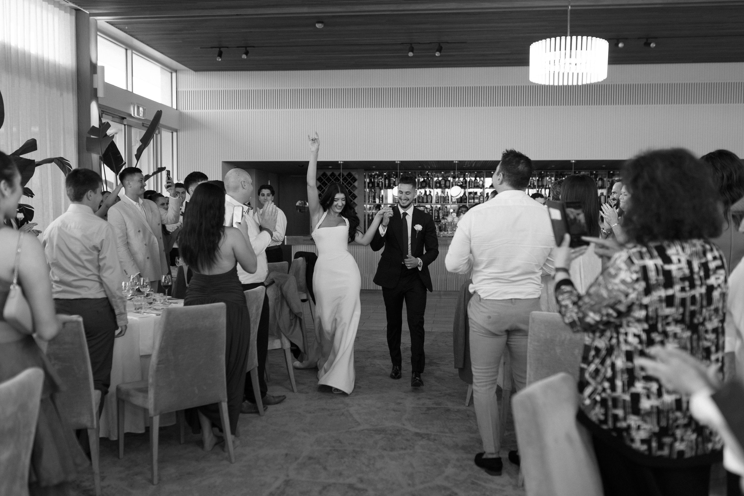 A black-and-white photo of a bride and groom walking into a reception area, holding hands and smiling, surrounded by guests taking photos and clapping.