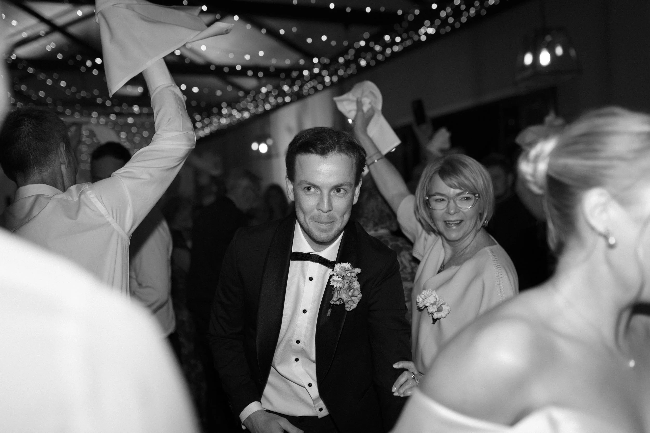 Black and white photo of people dancing at a celebration, with a man in a tuxedo in the center and women dancing around him; string lights hanging from the ceiling.
