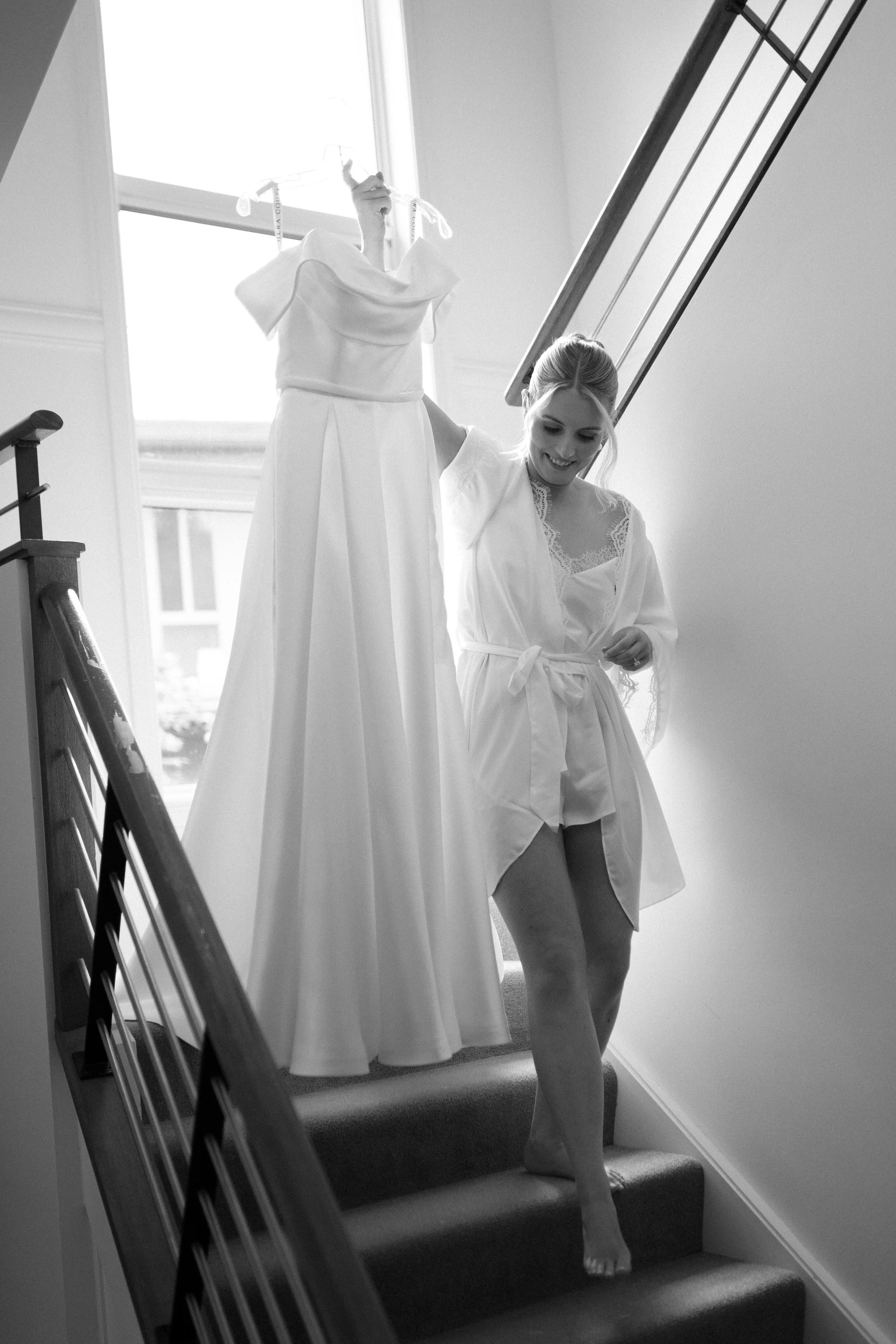 A woman in a white robe walking up a carpeted staircase, smiling, with a dress hanging on a hanger in front of her. The dress is likely for a wedding or special occasion.