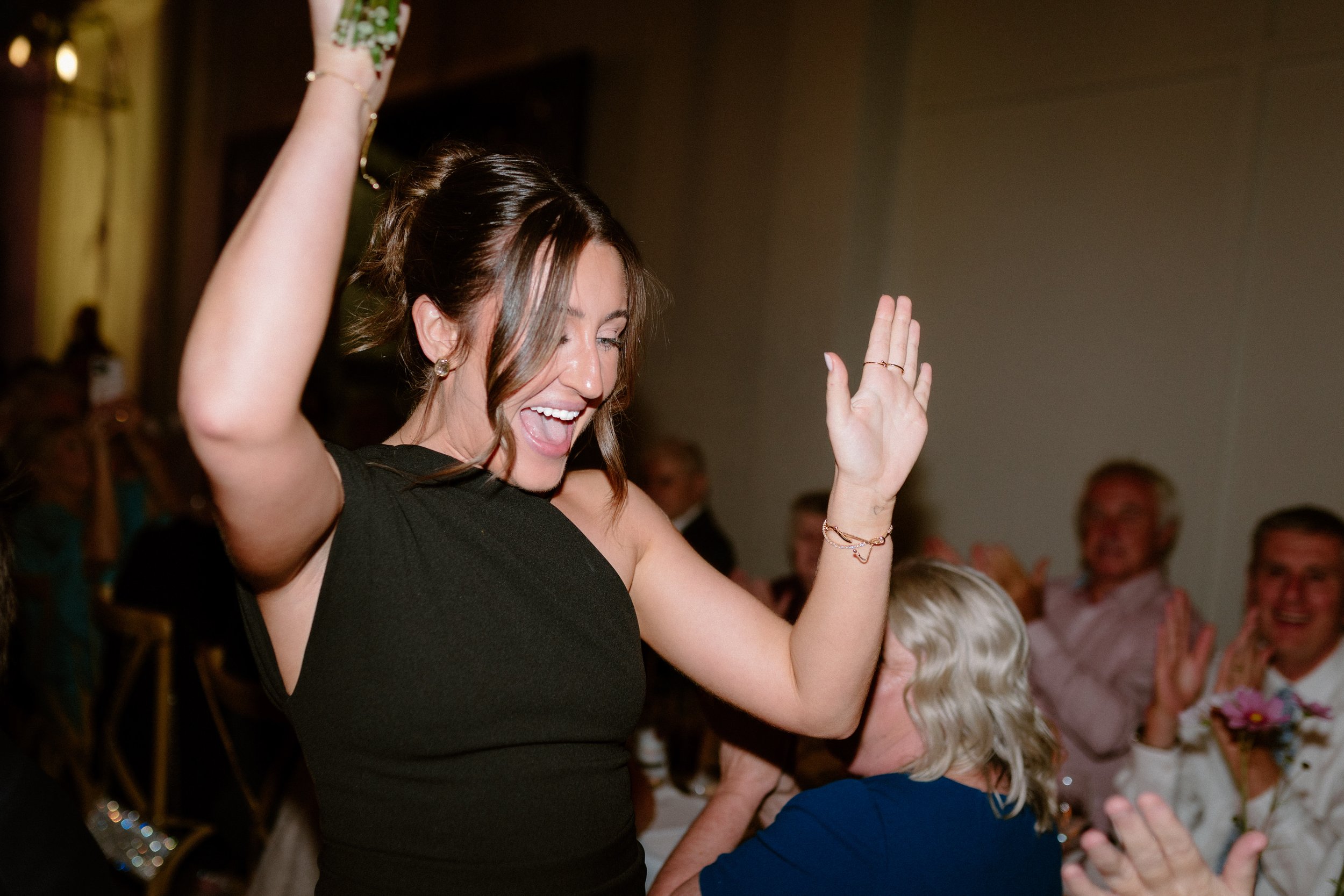 A woman with brown hair wearing a black dress dancing and smiling at an event, with other guests in the background.