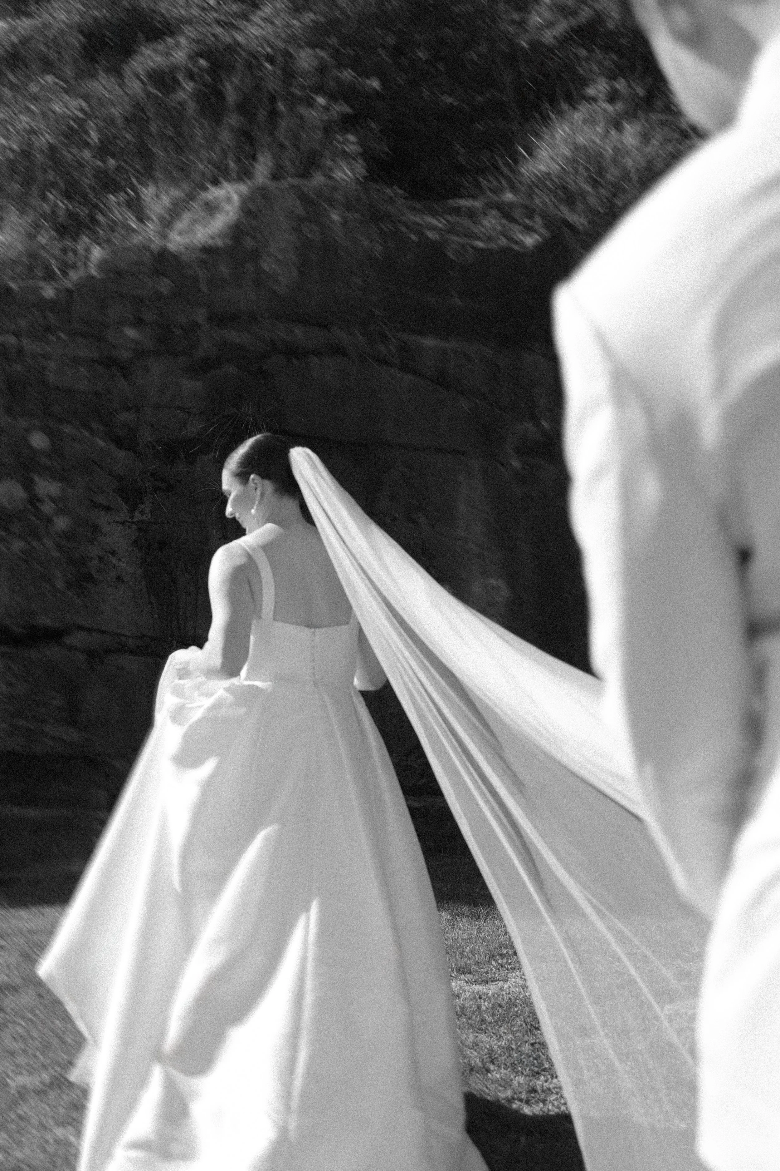 Black and white photo of a bride in a wedding dress with a long train, standing against a rocky background, looking over her shoulder.
