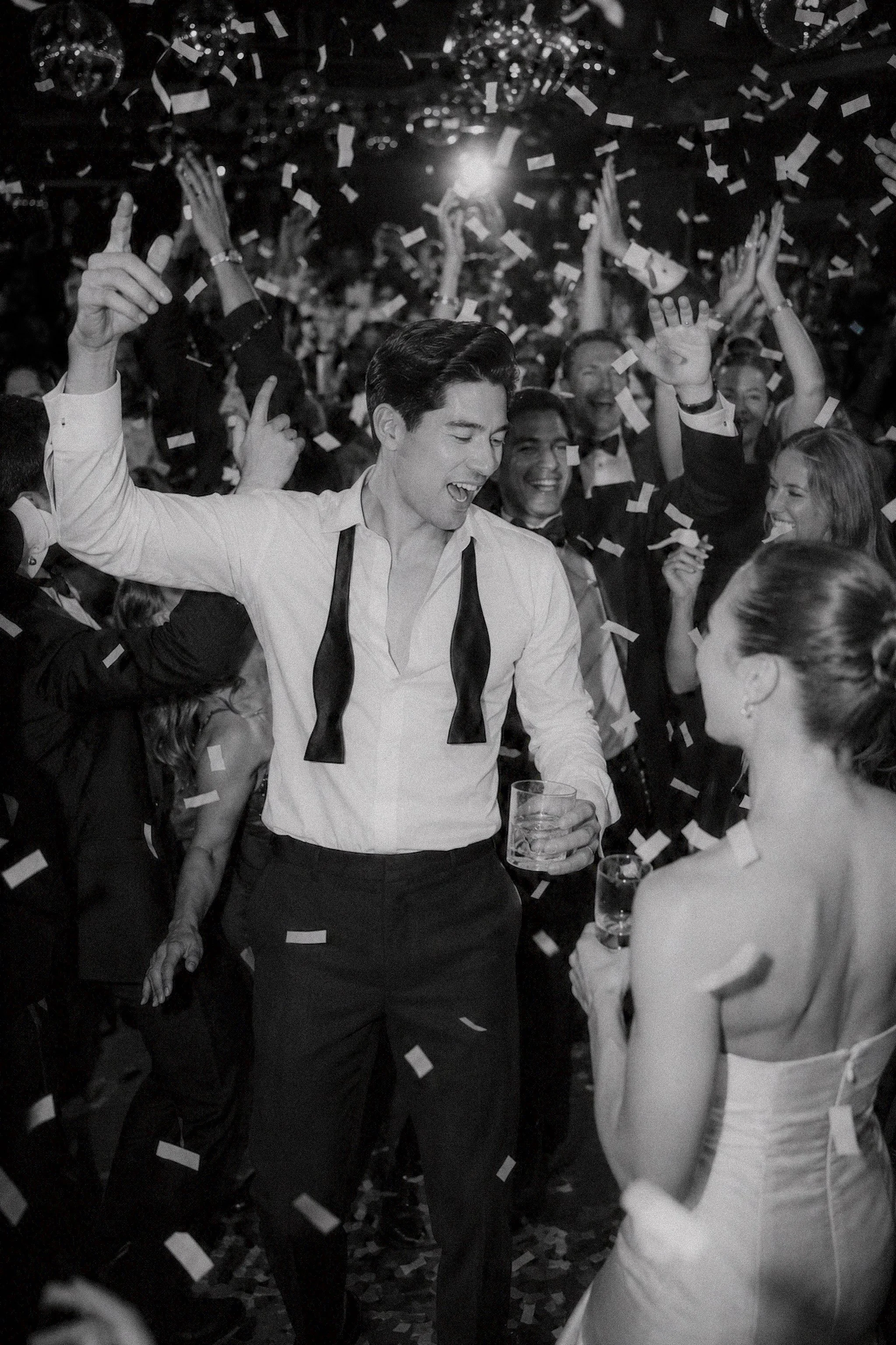 A young man in formal attire dancing at a lively celebration with people, confetti, and drinks, smiling and enjoying the moment.