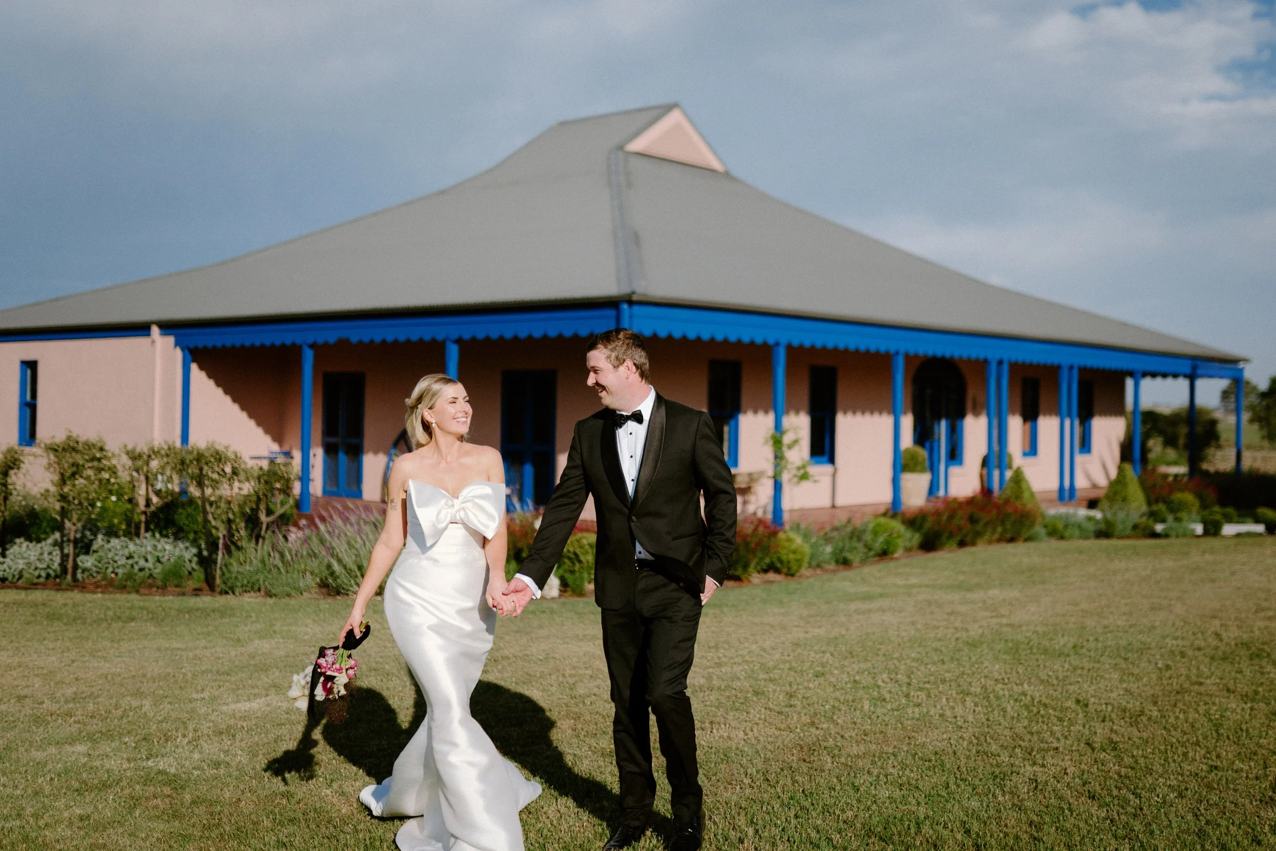 The Sienna, Hunter Valley Wedding Venue, Wedding Photos. A bride and groom holding hands and walking on grass in front of a pink building with blue accents, during their wedding day.