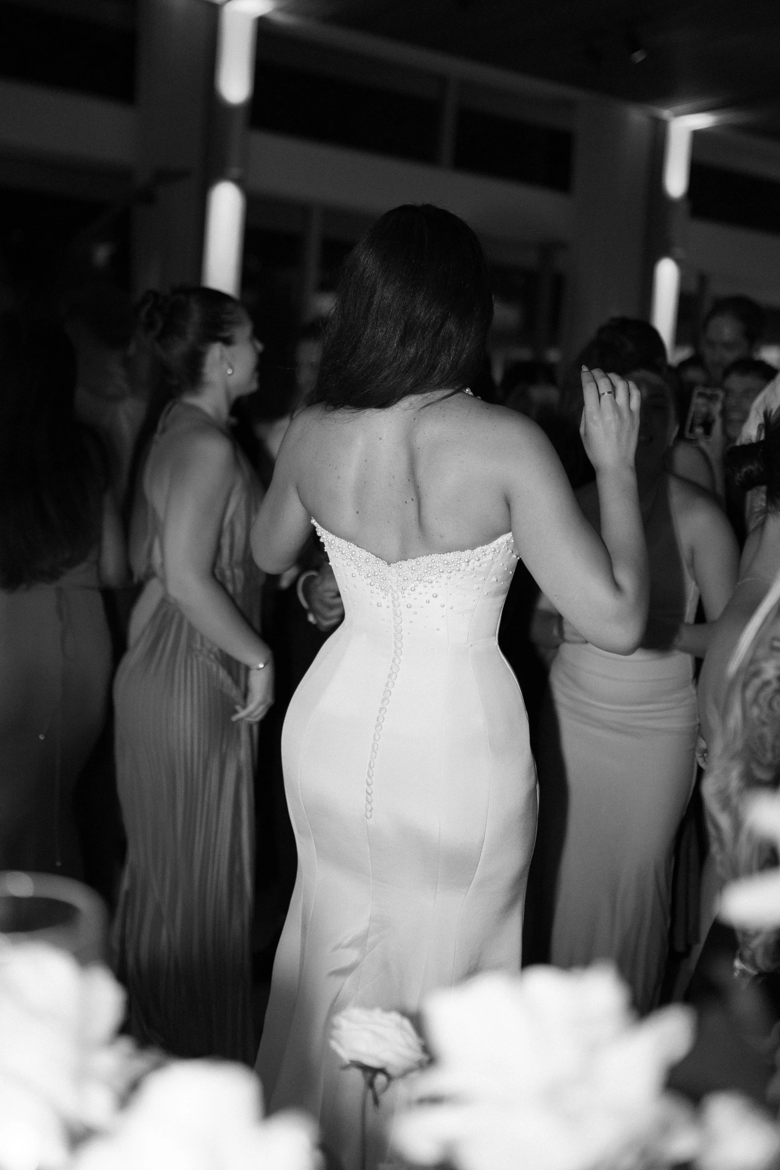 A woman in a strapless white wedding gown dancing at a party with other guests around her.