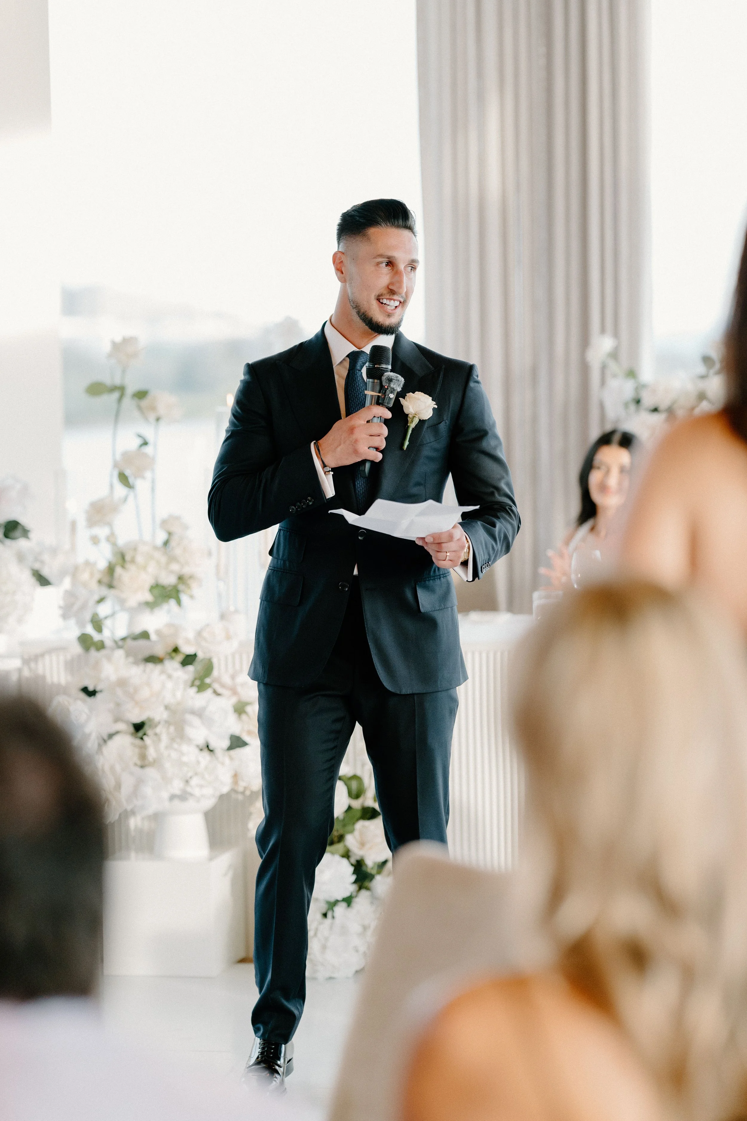 A man in a black suit giving a speech at a wedding reception, holding a paper and a microphone, with floral decorations in the background.