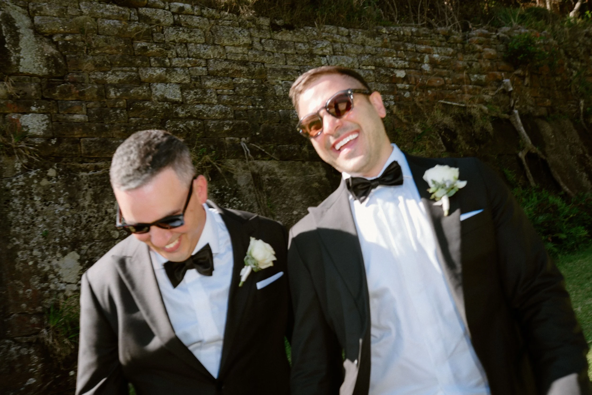 Two men in tuxedos and sunglasses smiling and looking down, standing outdoors near a stone wall.