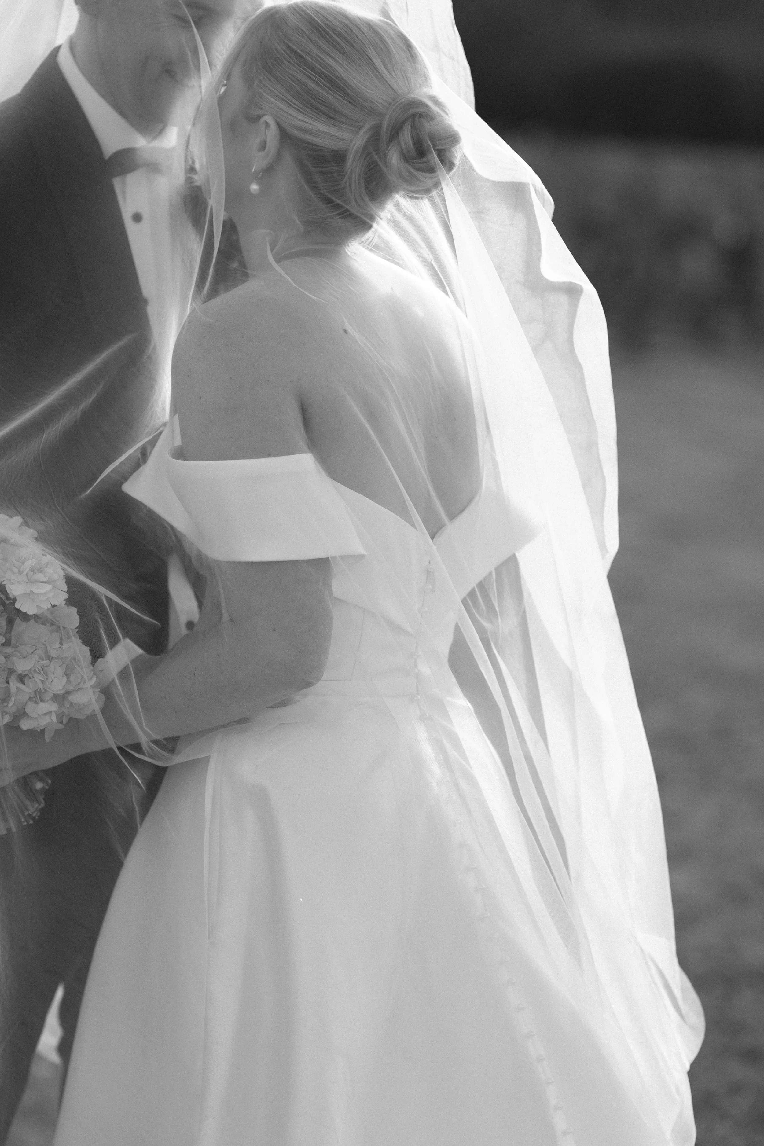 A bride with a veil and pearl earrings, holding a bouquet of flowers, leaning into a groom dressed in a tuxedo, during a wedding ceremony. Bimbadgen Palmers Lane Wedding, Hunter Valley