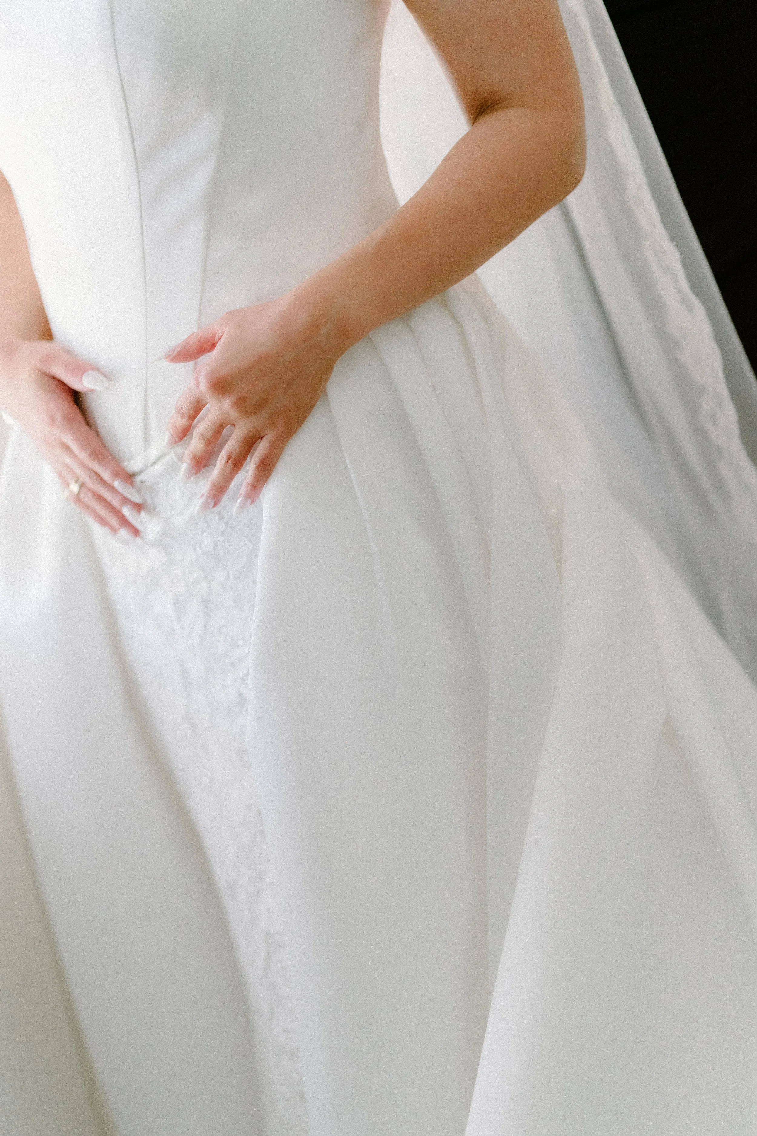 Close-up of hands of a bride in a white wedding gown with lace details, resting on her lap.