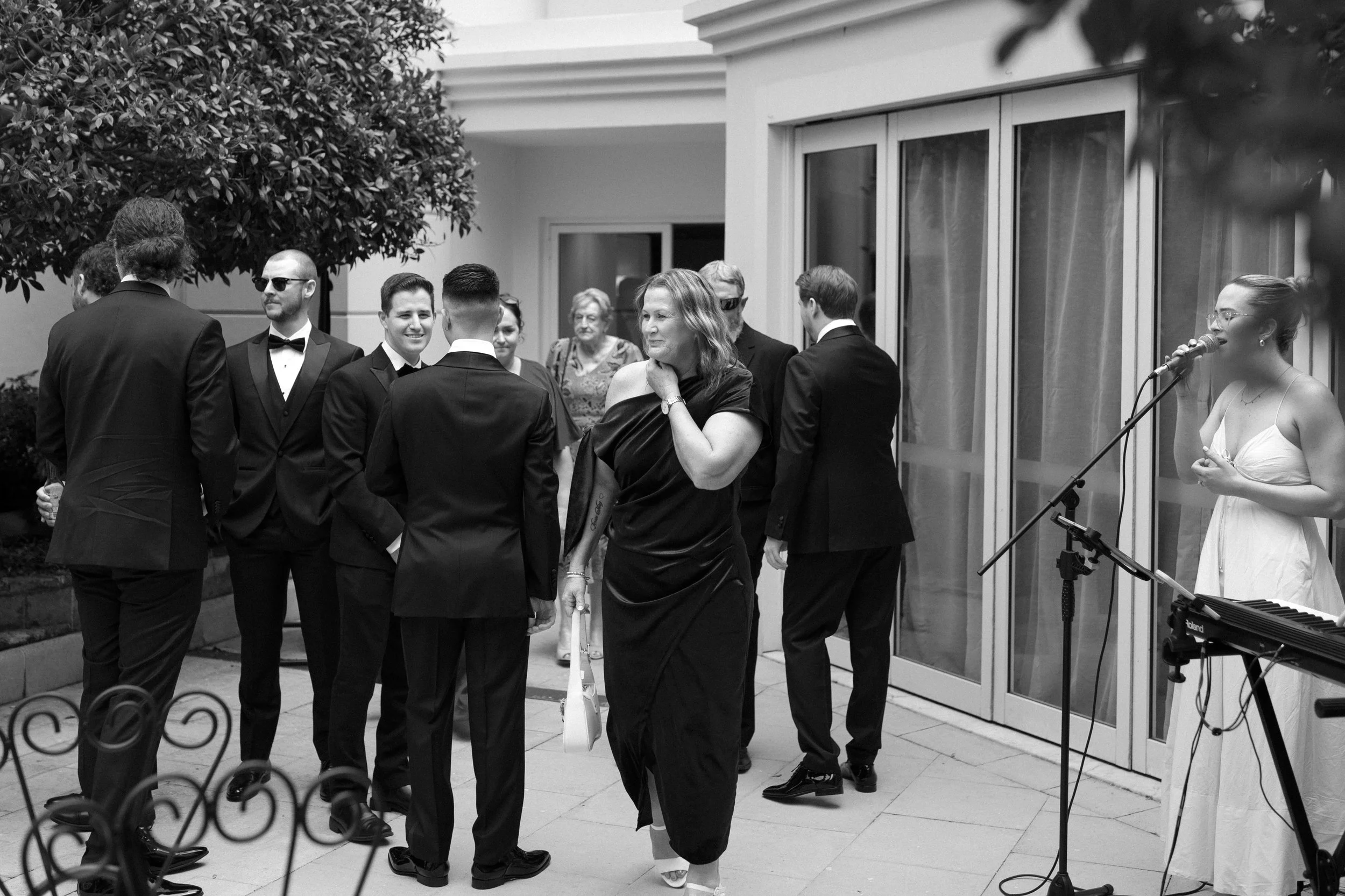 Guests in formal attire at an outdoor wedding reception, some chatting and a woman singing into a microphone near a keyboard.