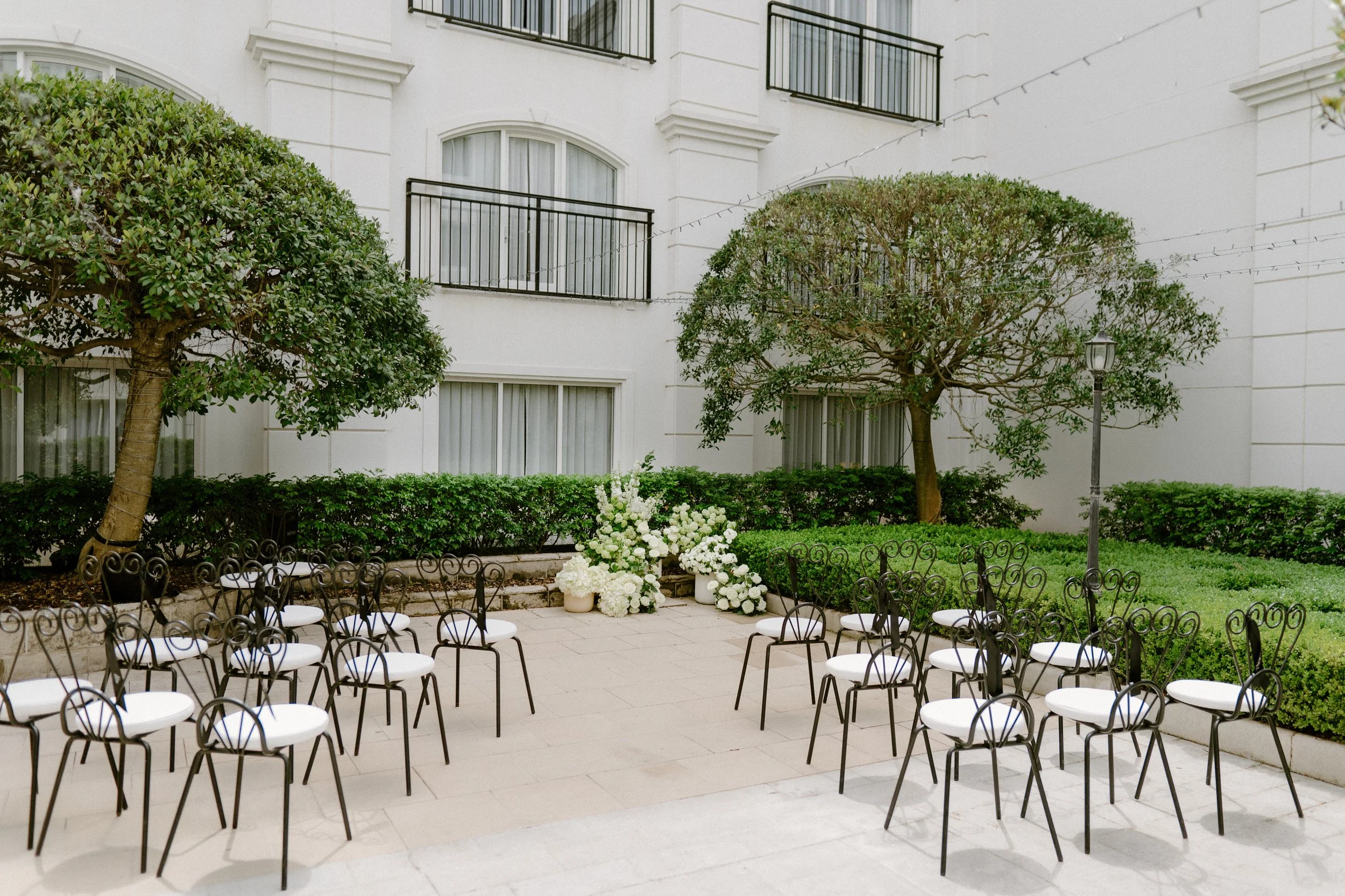 Outdoor wedding setup with white chairs arranged in a semi-circle, white floral arrangements, green bushes, trees, and string lights in a courtyard area.