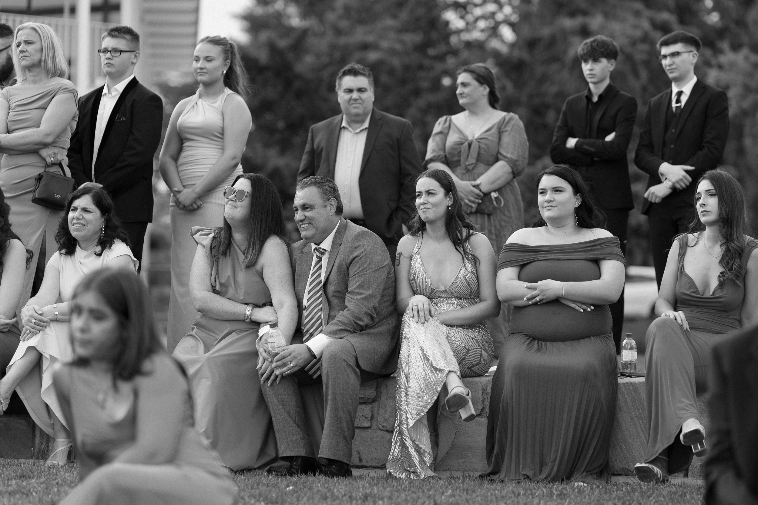A group of people sitting and standing outdoors during a formal event, with various expressions on their faces.