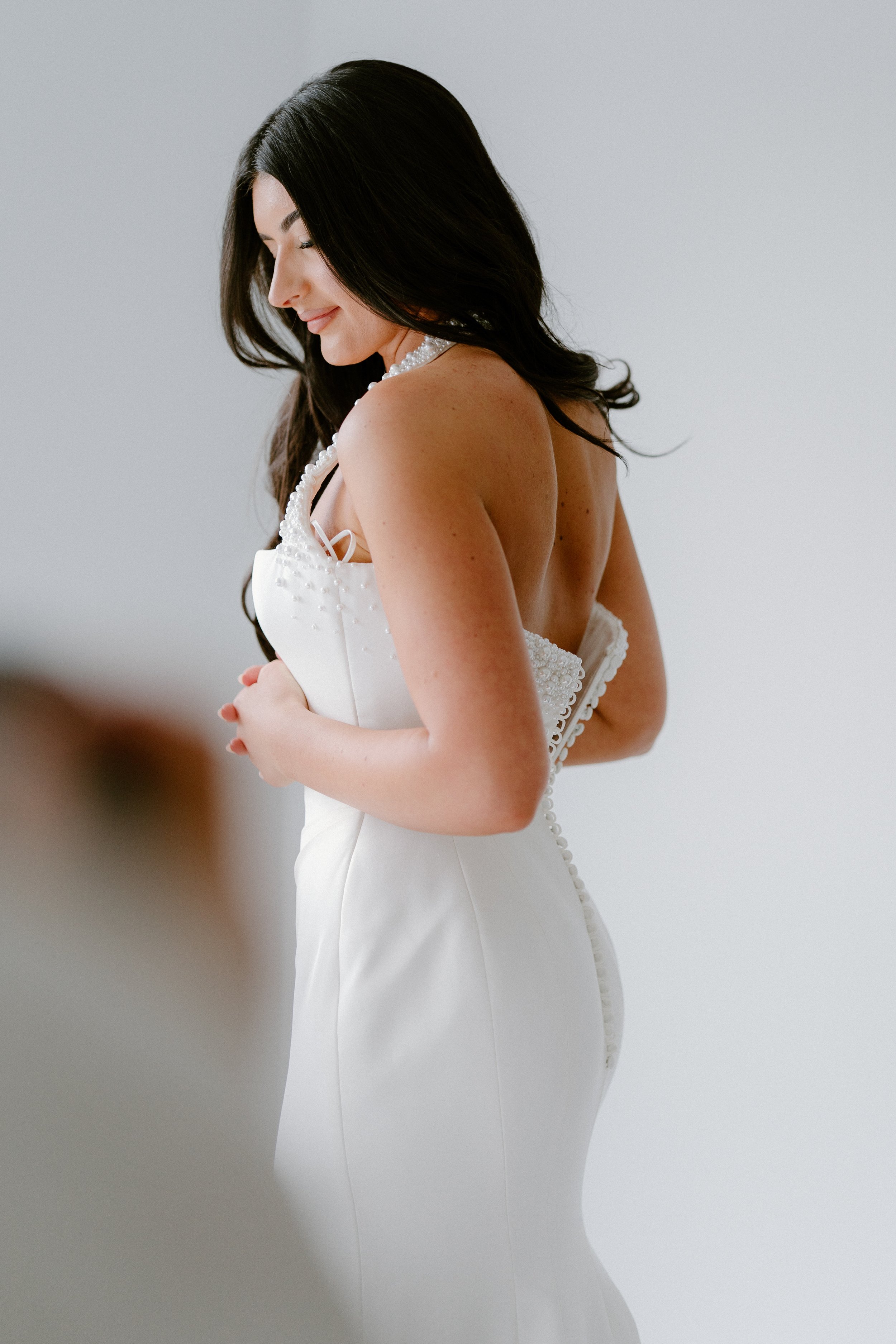 A woman in a white wedding dress with pearl embellishments and dark hair smiling, with a plain gray background.