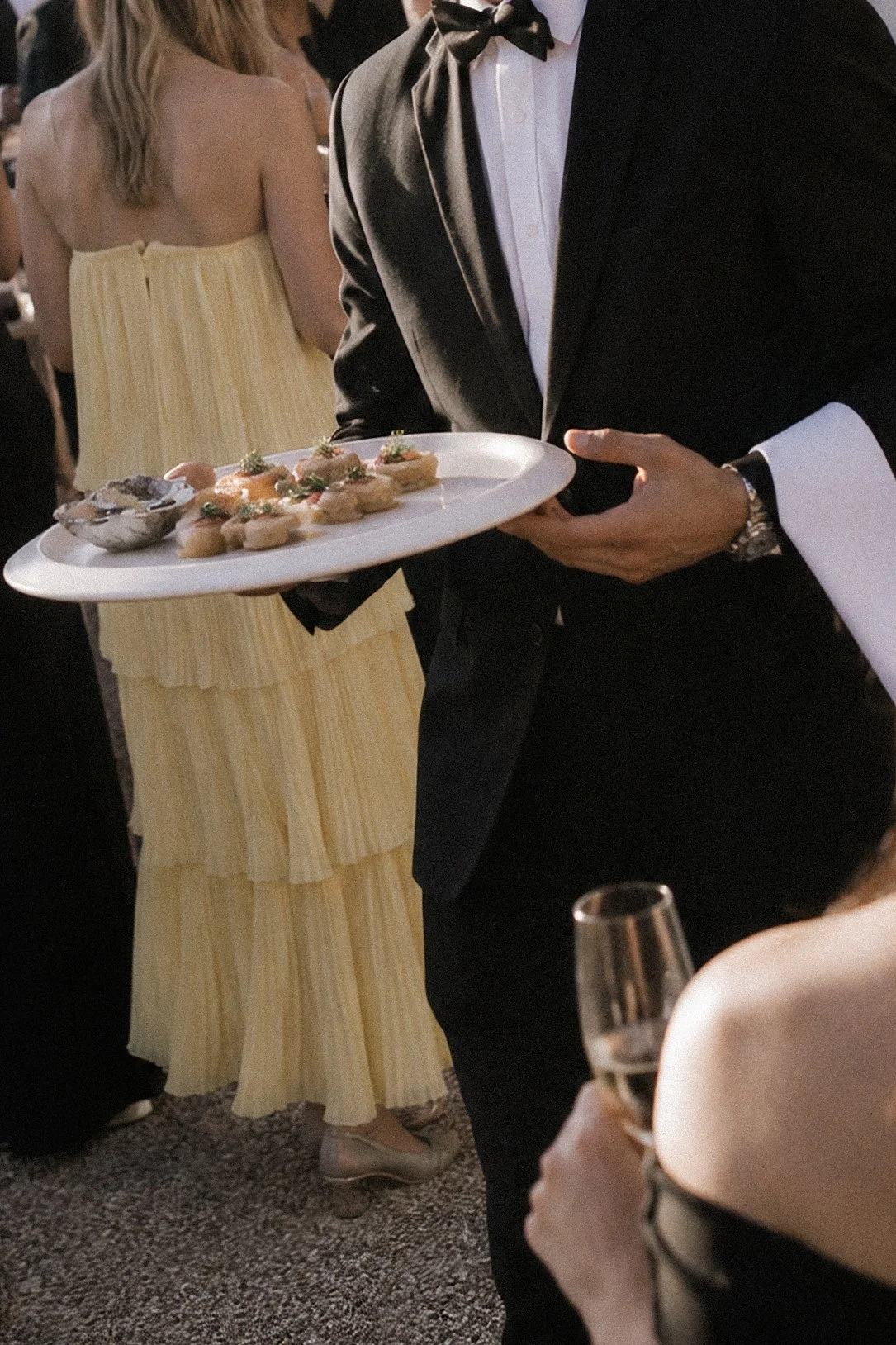 A man in a black tuxedo and bow tie holding a white tray with various appetizers and a dipping sauce at a formal event.