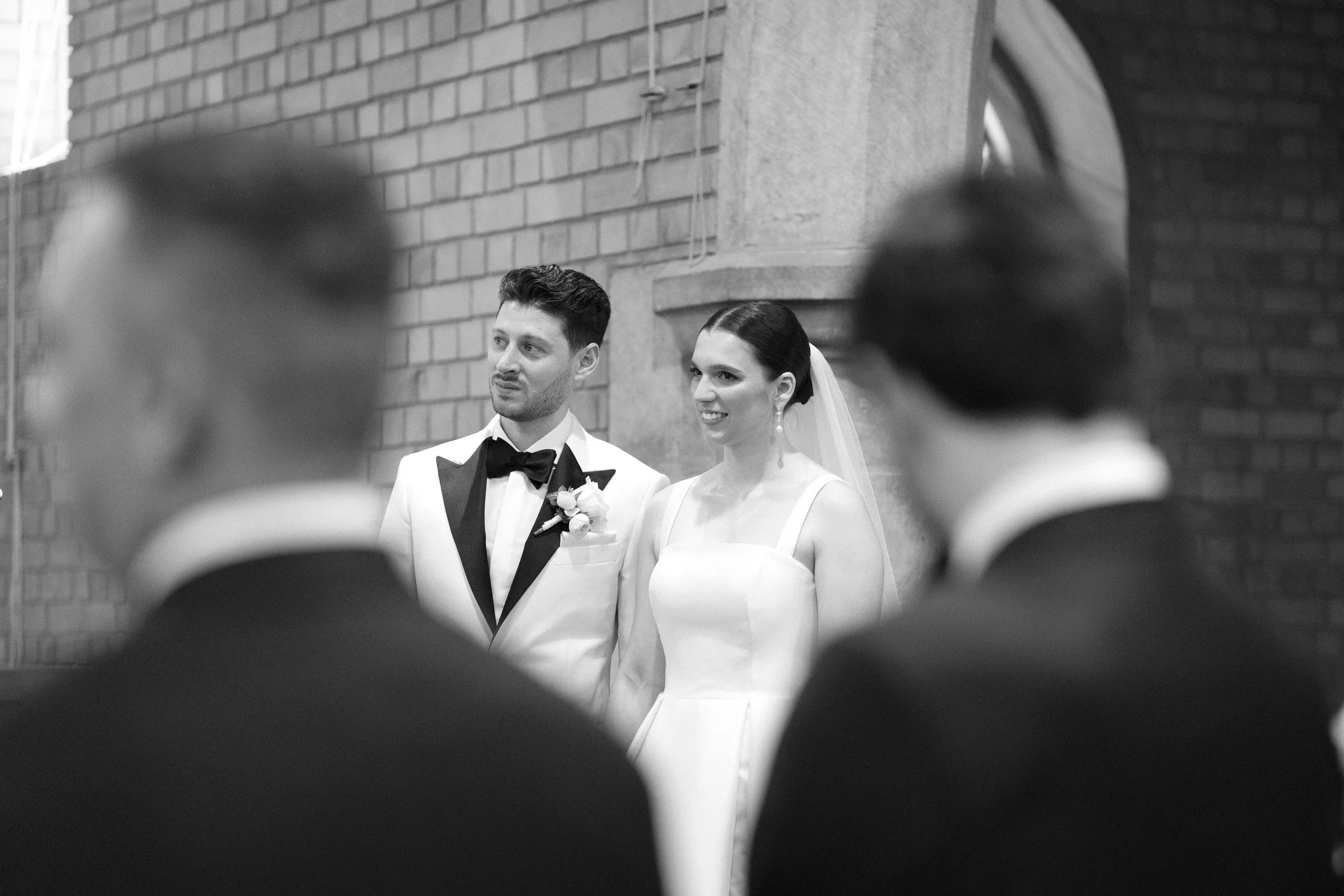 A black-and-white photo of a wedding ceremony, with the bride and groom standing in front of two blurred groomsmen. The bride is wearing a white dress with a veil, and the groom is in a tuxedo with a bow tie. They are inside a brick building.