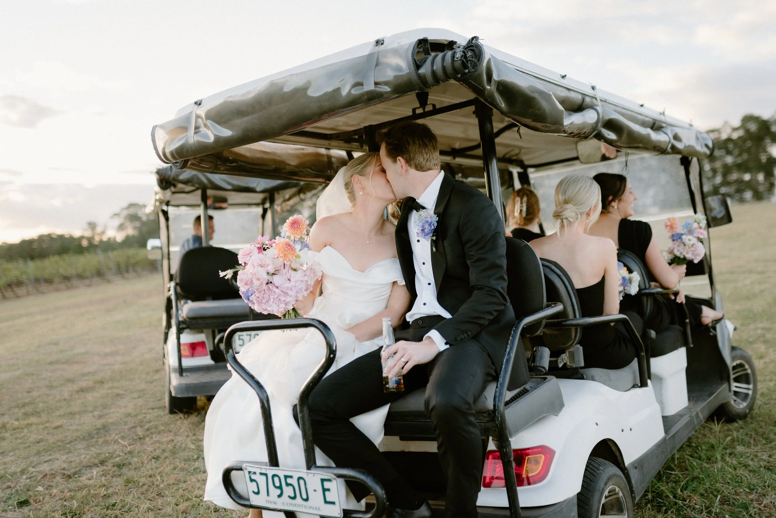 A newlywed couple sharing a kiss on a golf cart, with bridesmaids sitting behind them holding bouquets, at a wedding event outdoors. Bimbadgen Palmers Lane Wedding, Hunter Valley