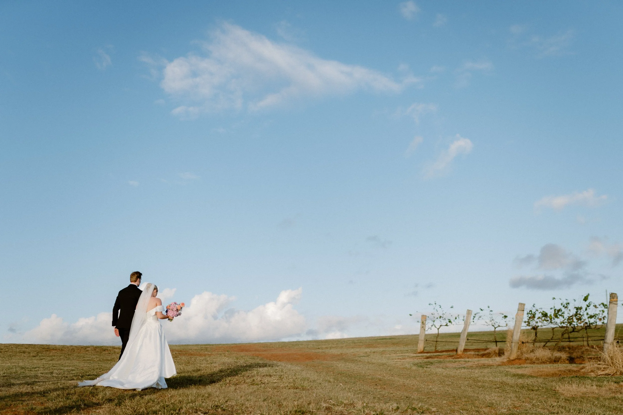 A bride and groom walking together on a grassy field under a blue sky with clouds, with the groom in a black suit and the bride in a white wedding dress holding a bouquet of flowers. Bimbadgen Palmers Lane Wedding, Hunter Valley