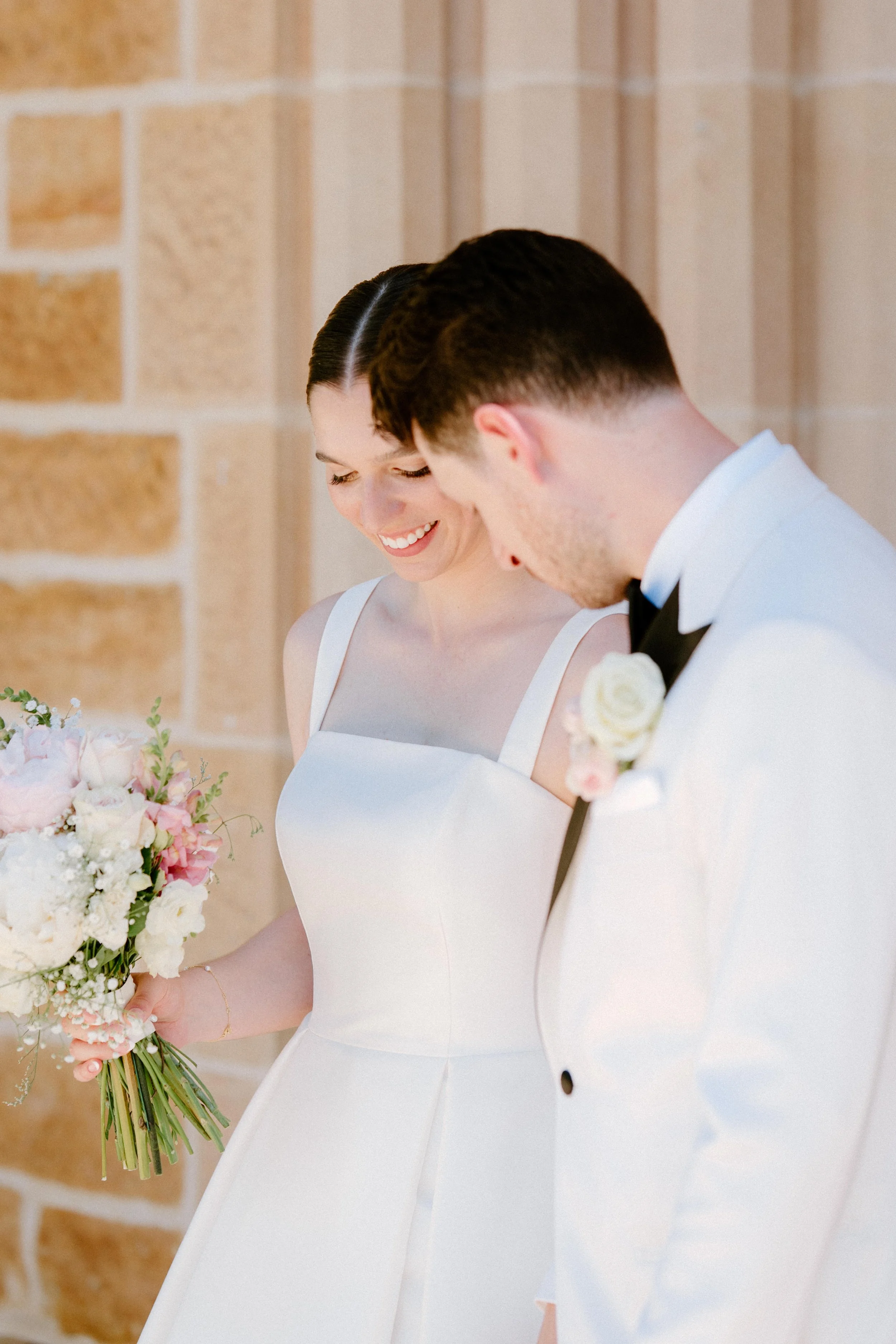 A bride and groom smiling and looking down, standing with a floral bouquet, against a brick wall background.