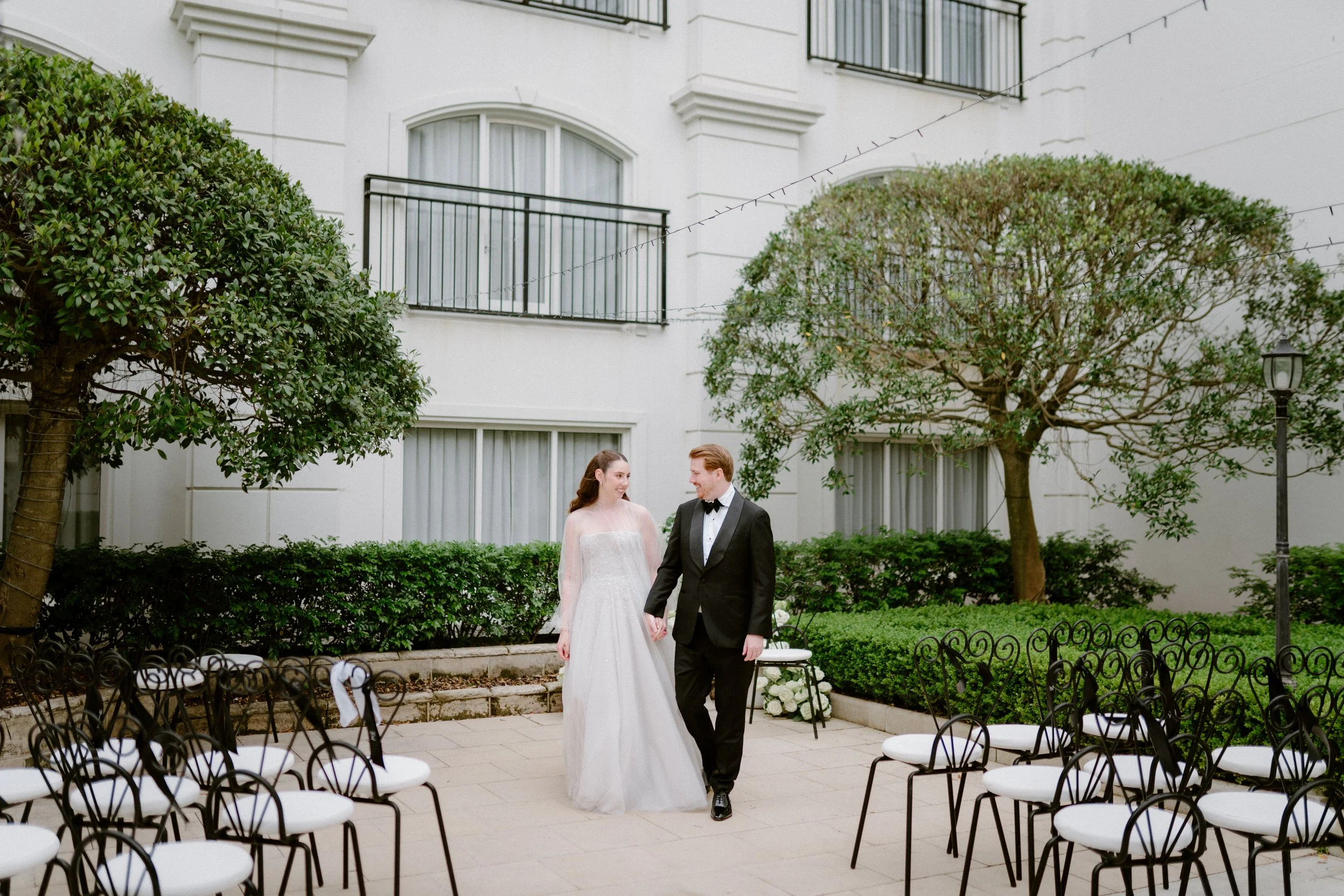 A bride and groom holding hands and smiling at each other during an outdoor wedding ceremony in a courtyard with chairs, trees, and shrubbery.