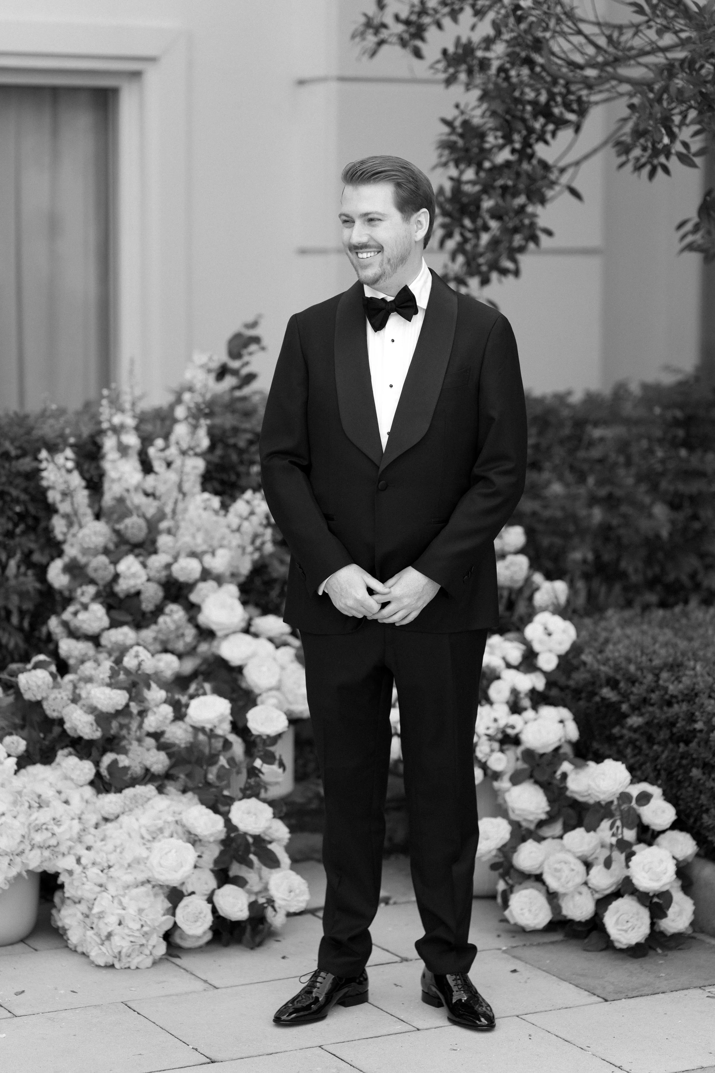 A man in a tuxedo smiling, standing outdoors near flower arrangements.