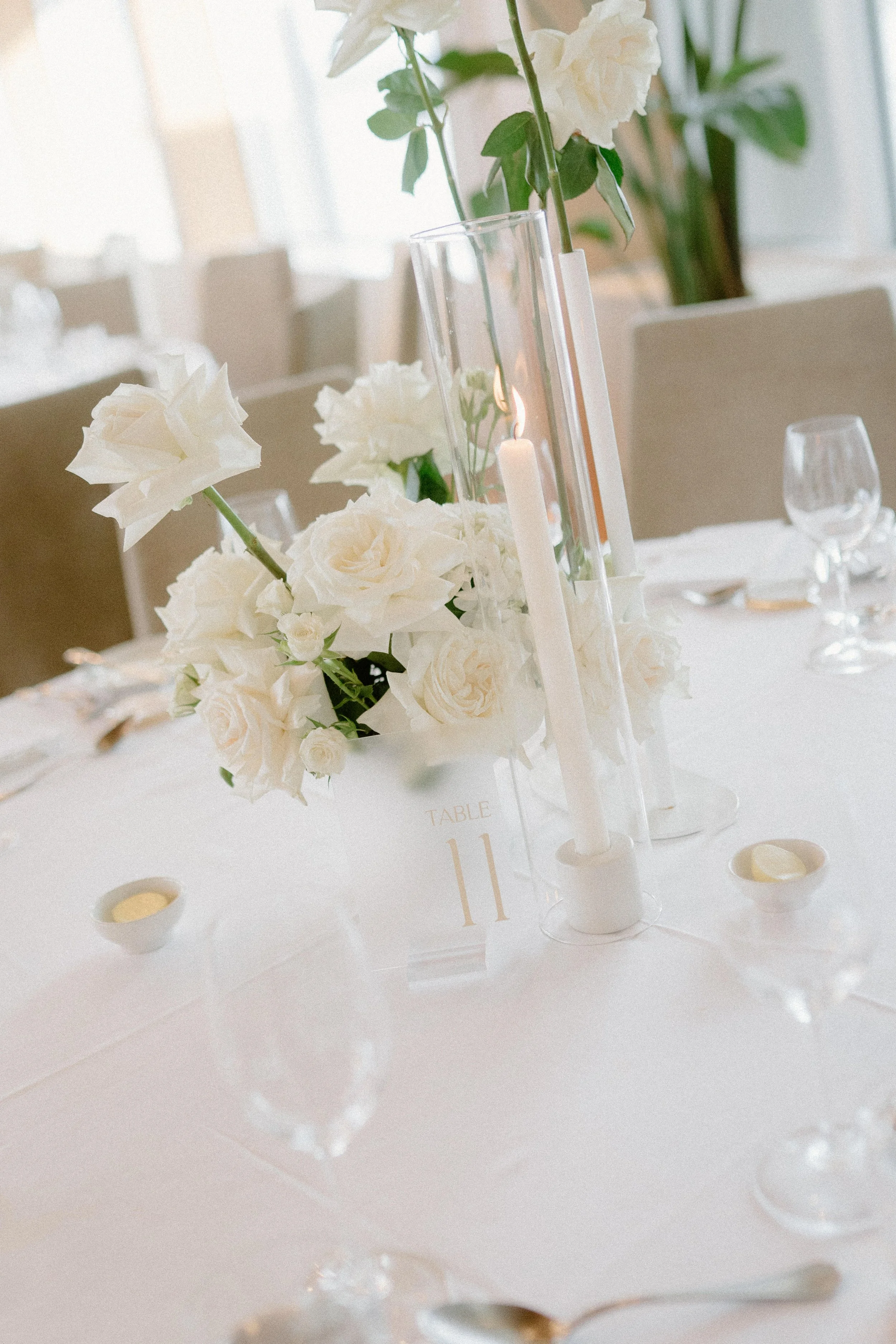 Elegant table centerpiece with white roses, white flowers, tall white candles in glass holders, and a table number placard that reads 'Table 11,' set on a white tablecloth in a decorated dining area.