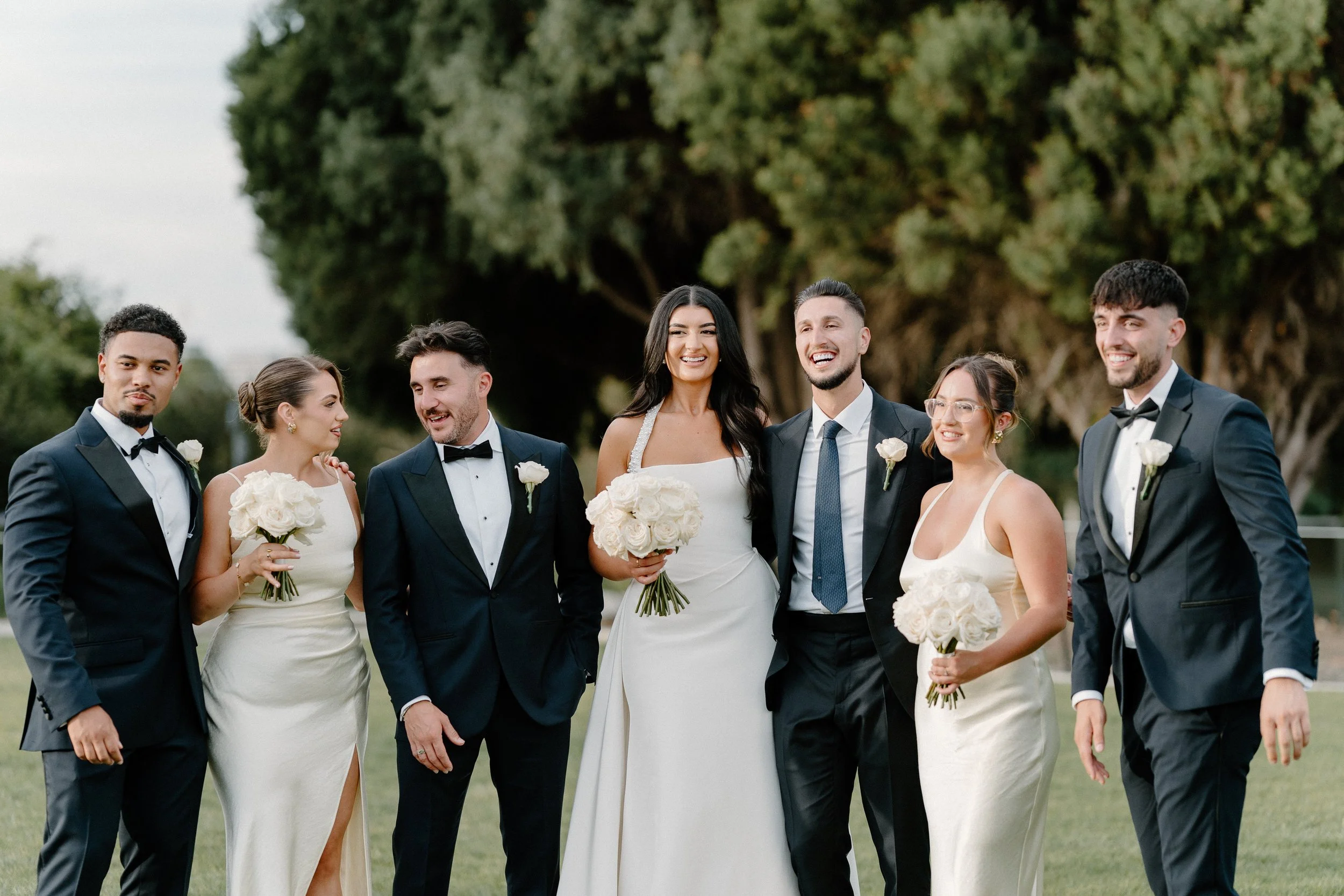 Group of seven people in wedding attire smiling outdoors with trees in background, celebrating a wedding.