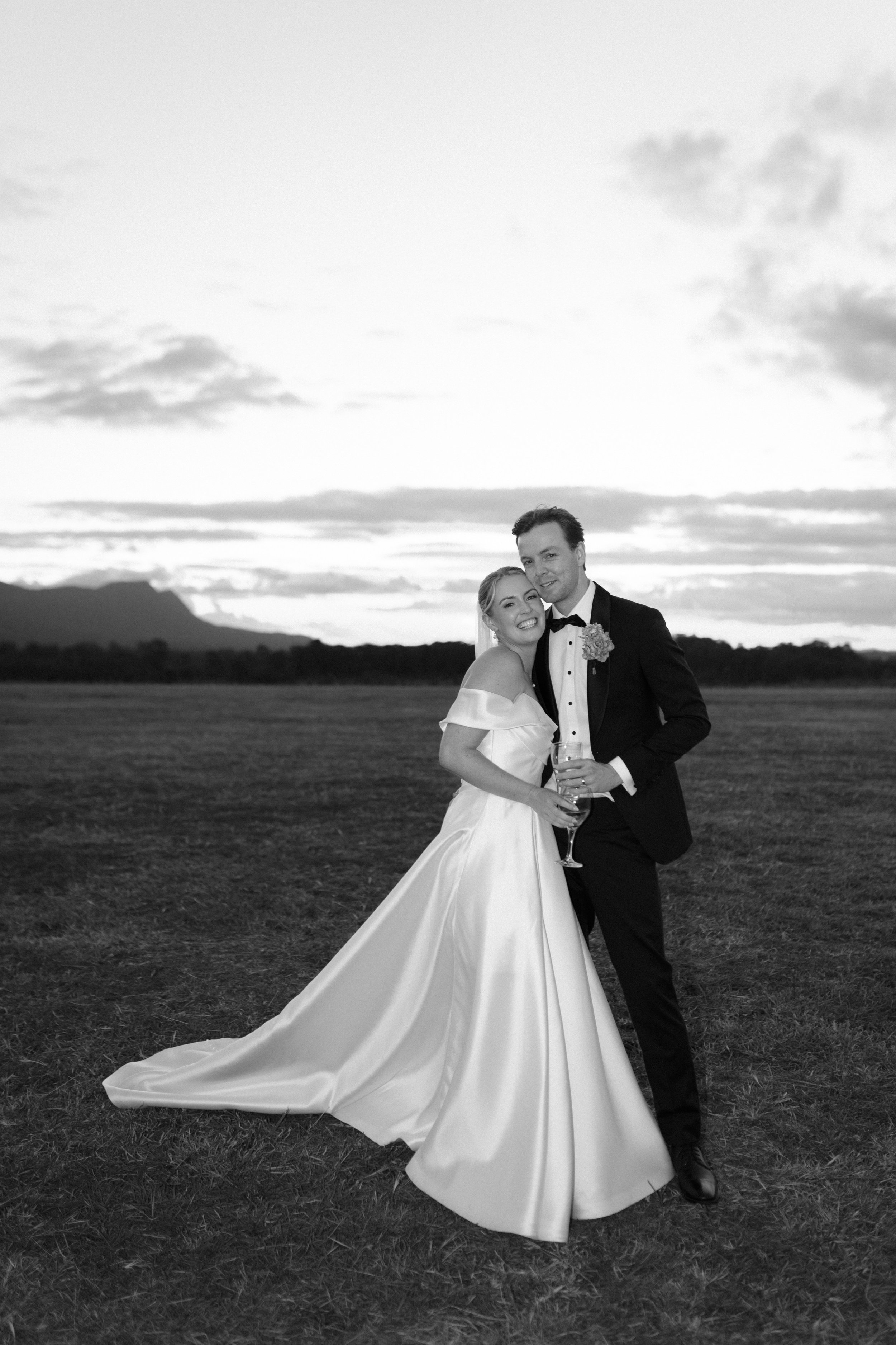 A black-and-white photo of a bride and groom smiling and posing outdoors at sunset, with the bride wearing an off-shoulder wedding gown and holding a glass, and the groom dressed in a suit with a bow tie, standing on grass with mountains and a cloudy