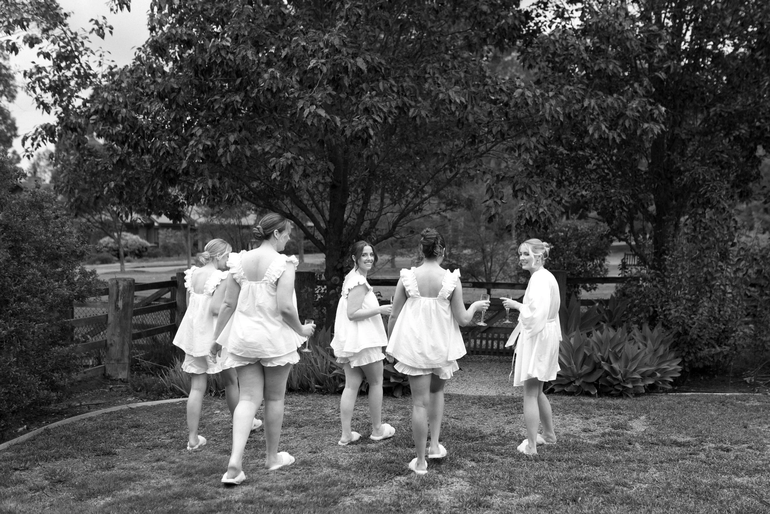 A group of women in white dresses standing outdoors near a large tree, holding glasses of champagne, engaging in conversation.