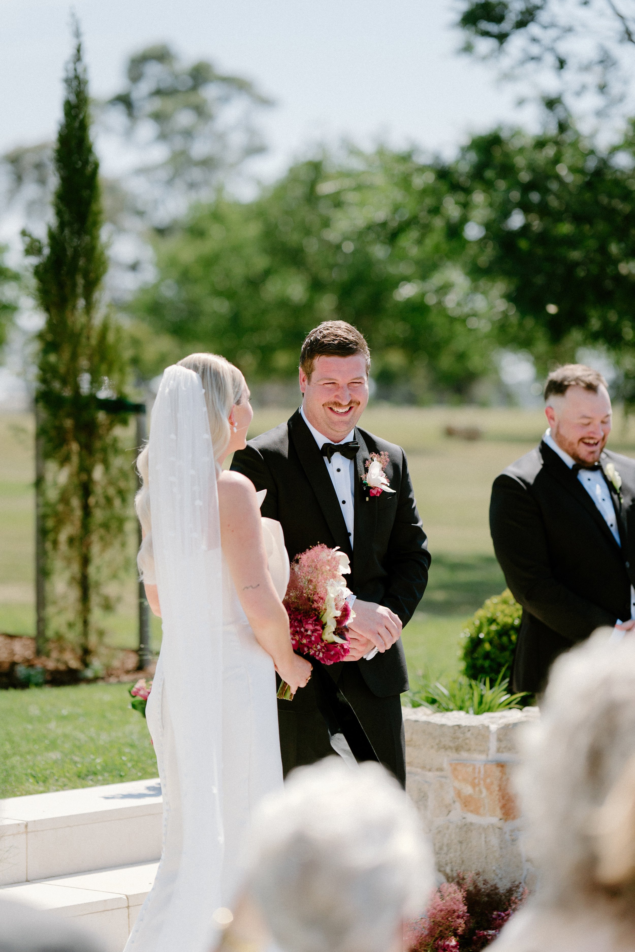 A bride and groom standing at an outdoor wedding ceremony, smiling and holding a bouquet of pink and white flowers, with a man in a tuxedo on the right, and trees in the background.