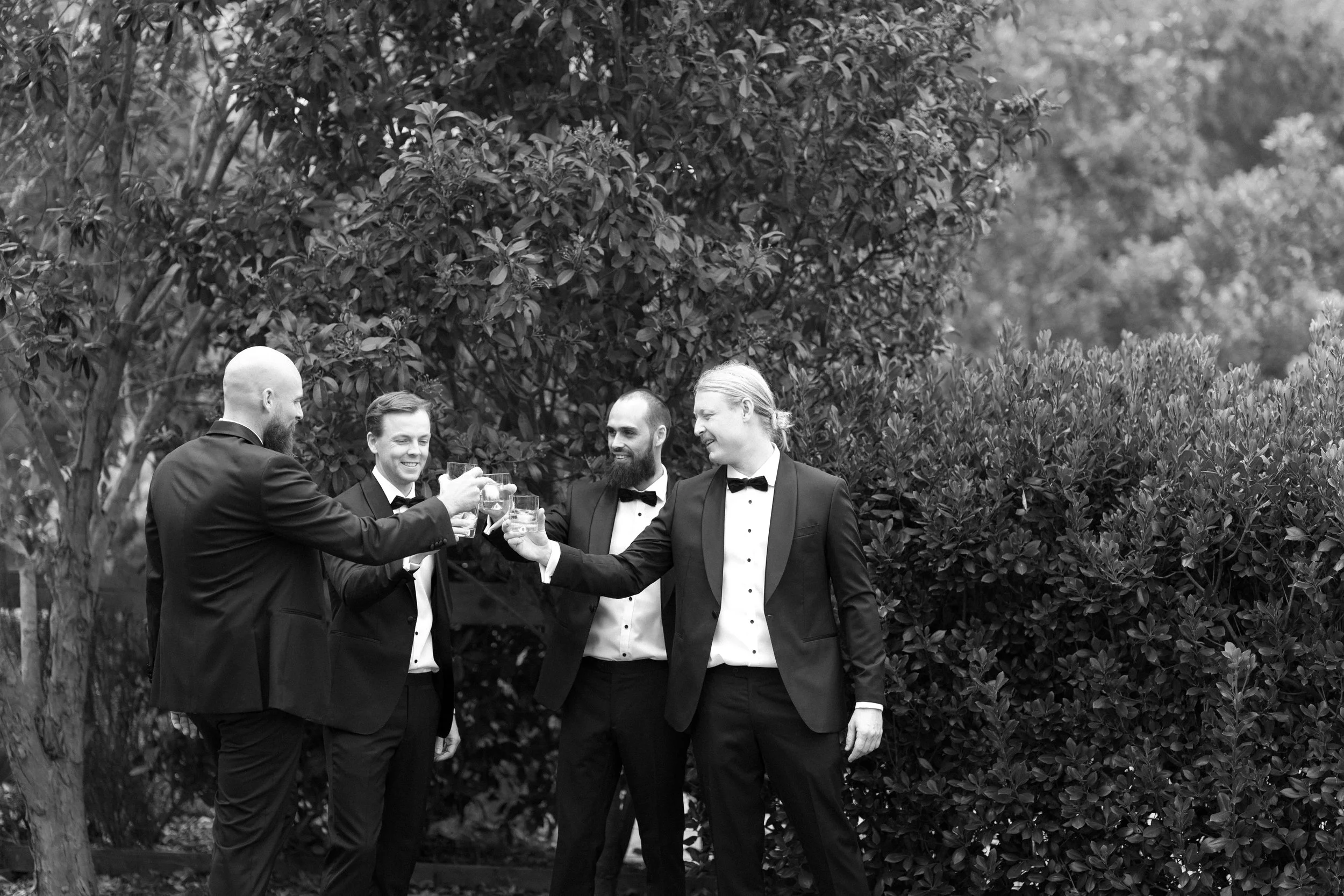 Four men in tuxedos raising glasses in a toast outdoors in front of bushes.