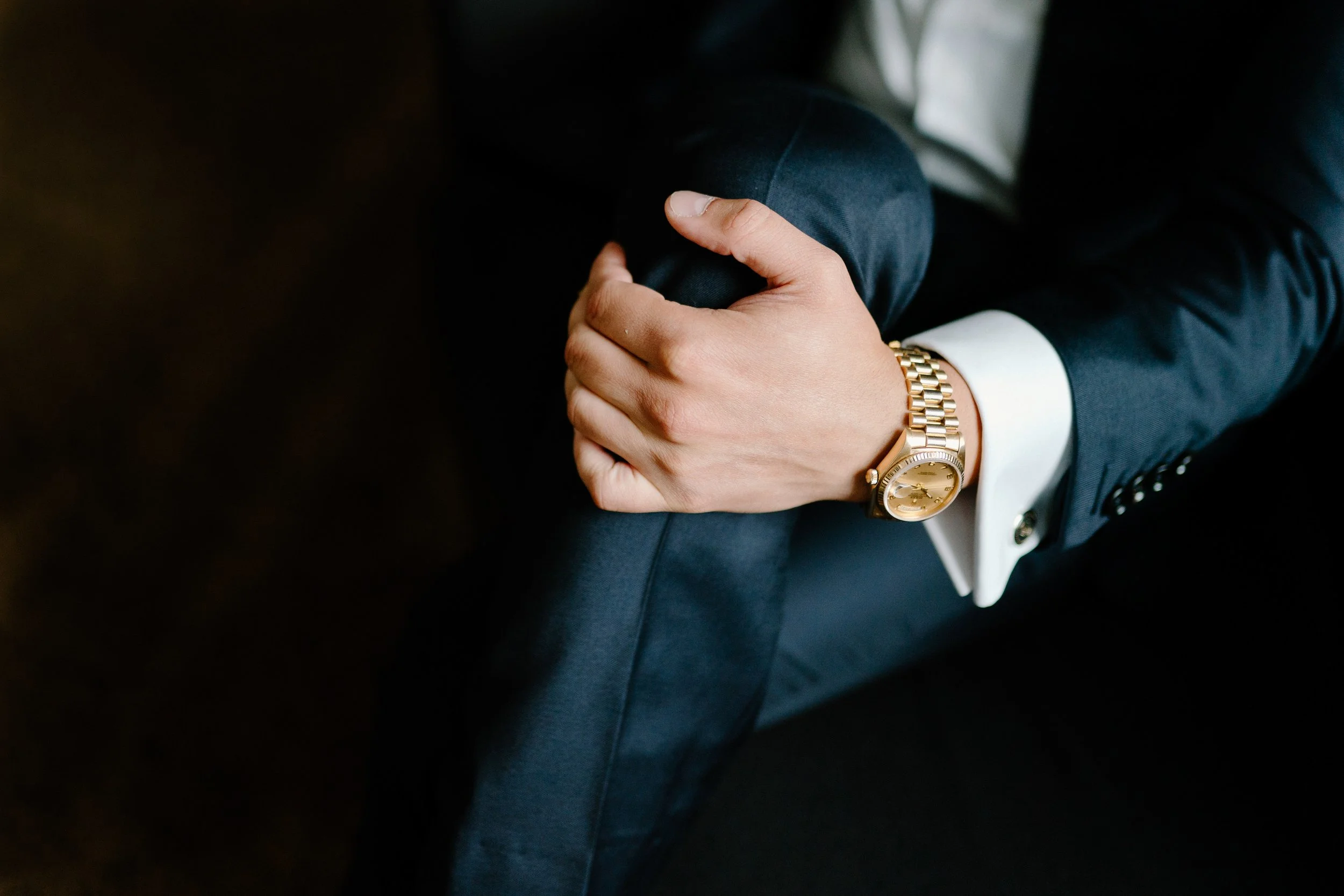 A person dressed in a dark suit with a white shirt sitting with one leg crossed over the other. They are wearing a gold watch on their left wrist. Their left hand is resting on their knee.