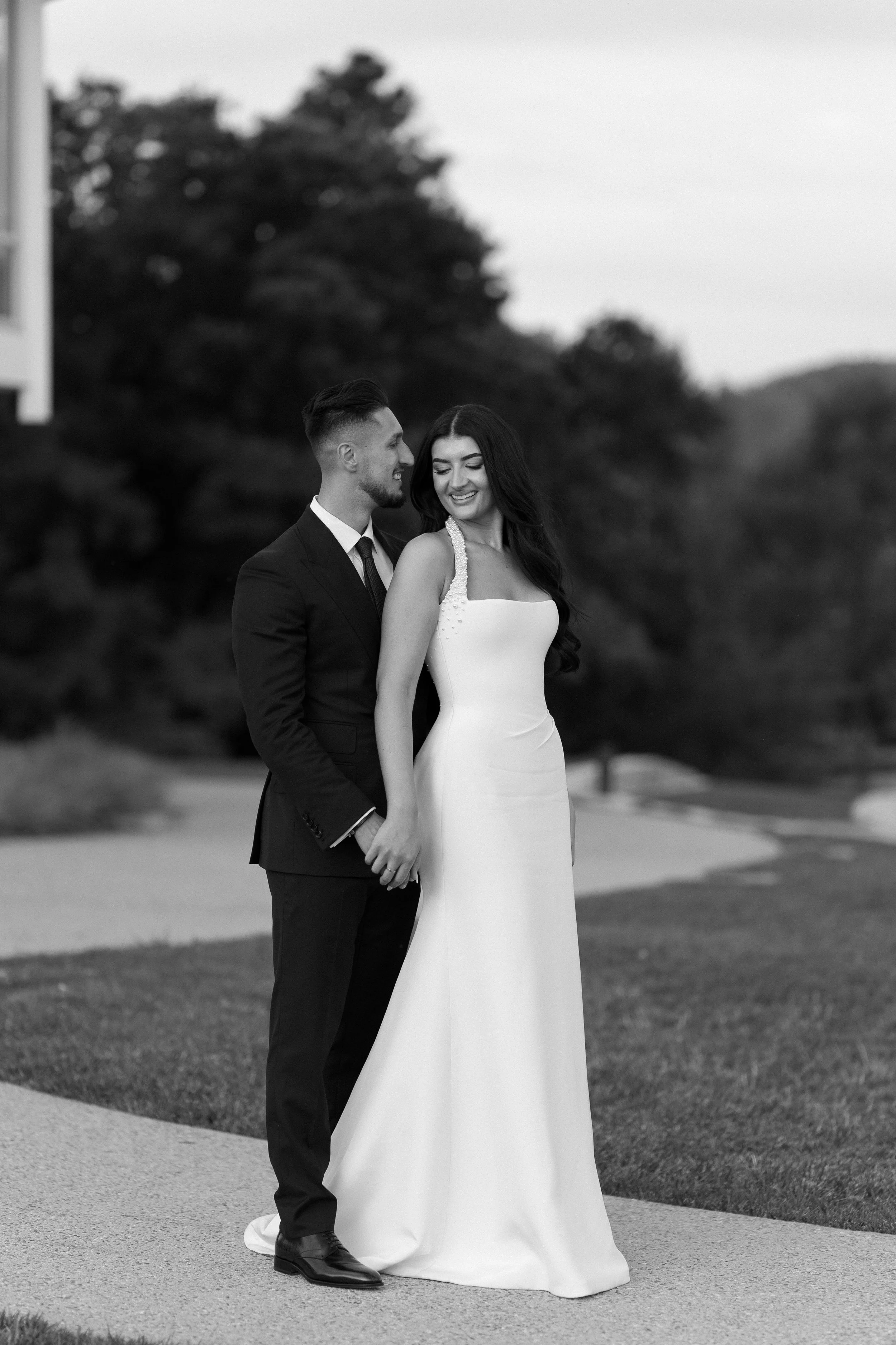 A black-and-white photo of a newlywed couple holding hands outdoors, smiling and looking at each other, with trees in the background.