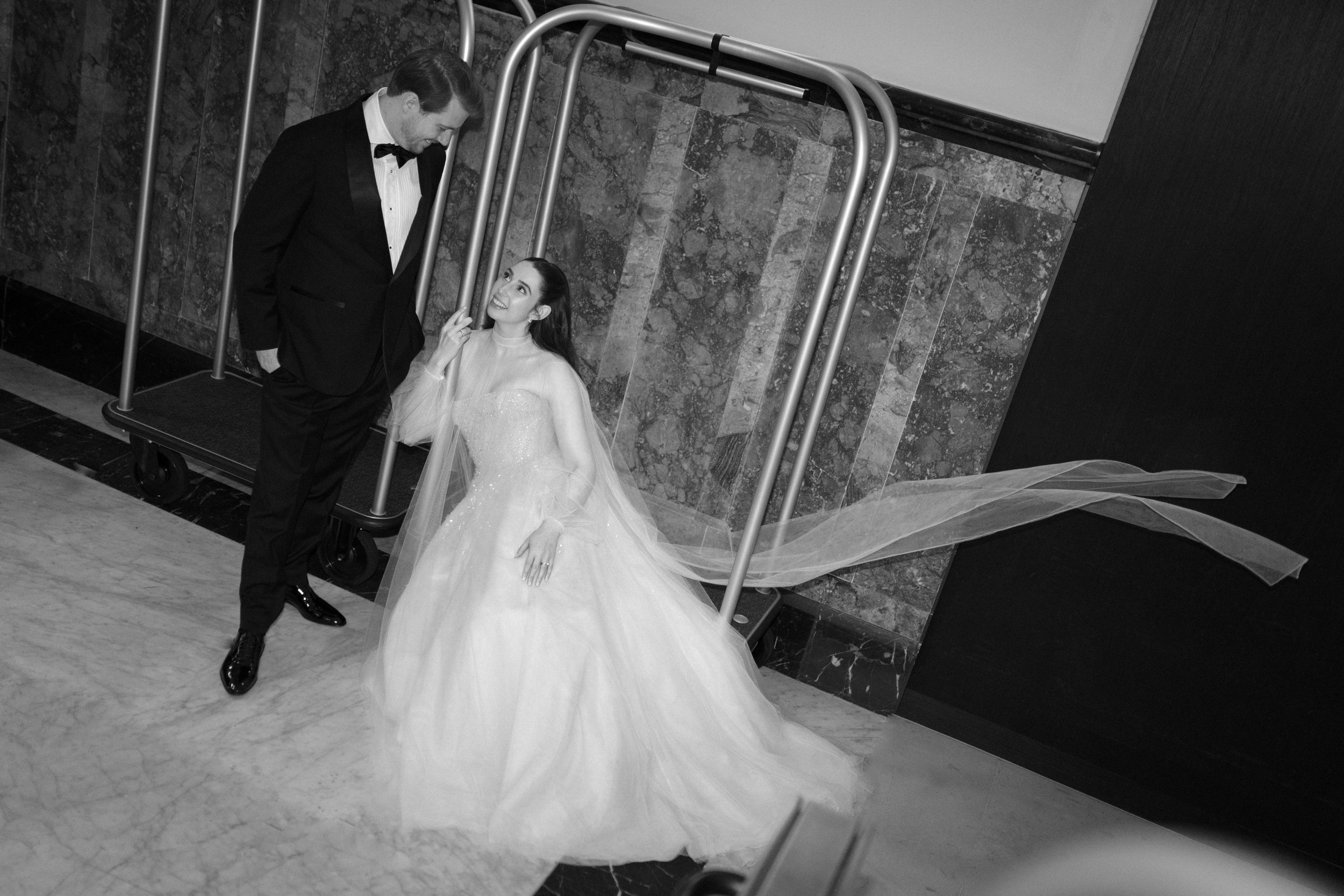 A woman in a wedding dress and a man in a tuxedo are smiling and looking at each other, standing on a marble floor near a luggage cart against a stone and dark-colored wall.