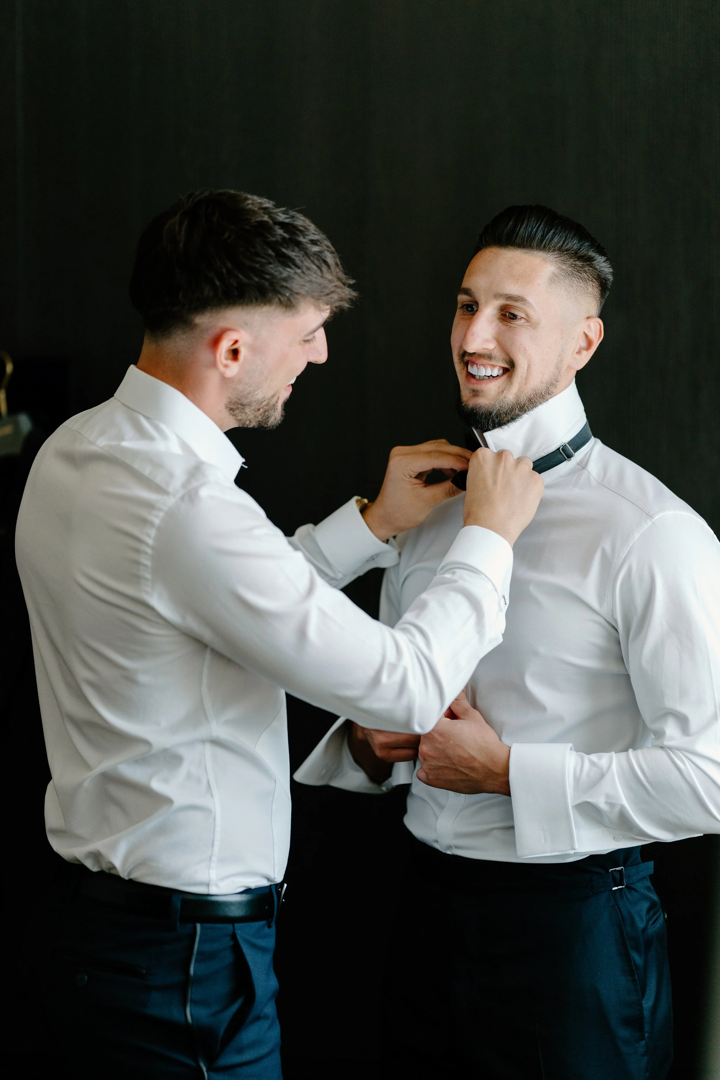 Two men in white dress shirts; one is helping the other adjust a black bow tie.