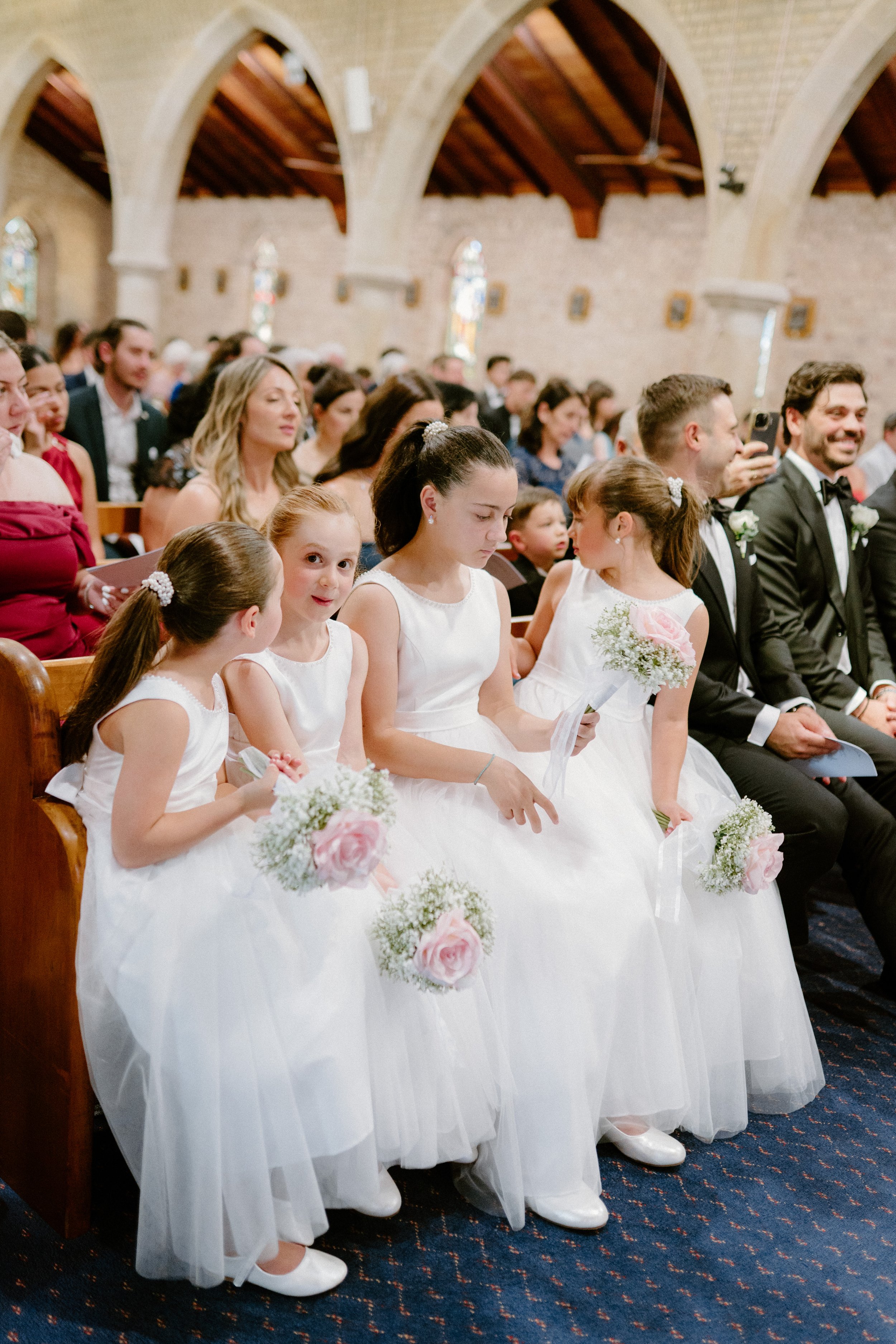 A group of young girls in white flower girl dresses holding bouquets seated in a church during a wedding ceremony, with other guests seated behind them.