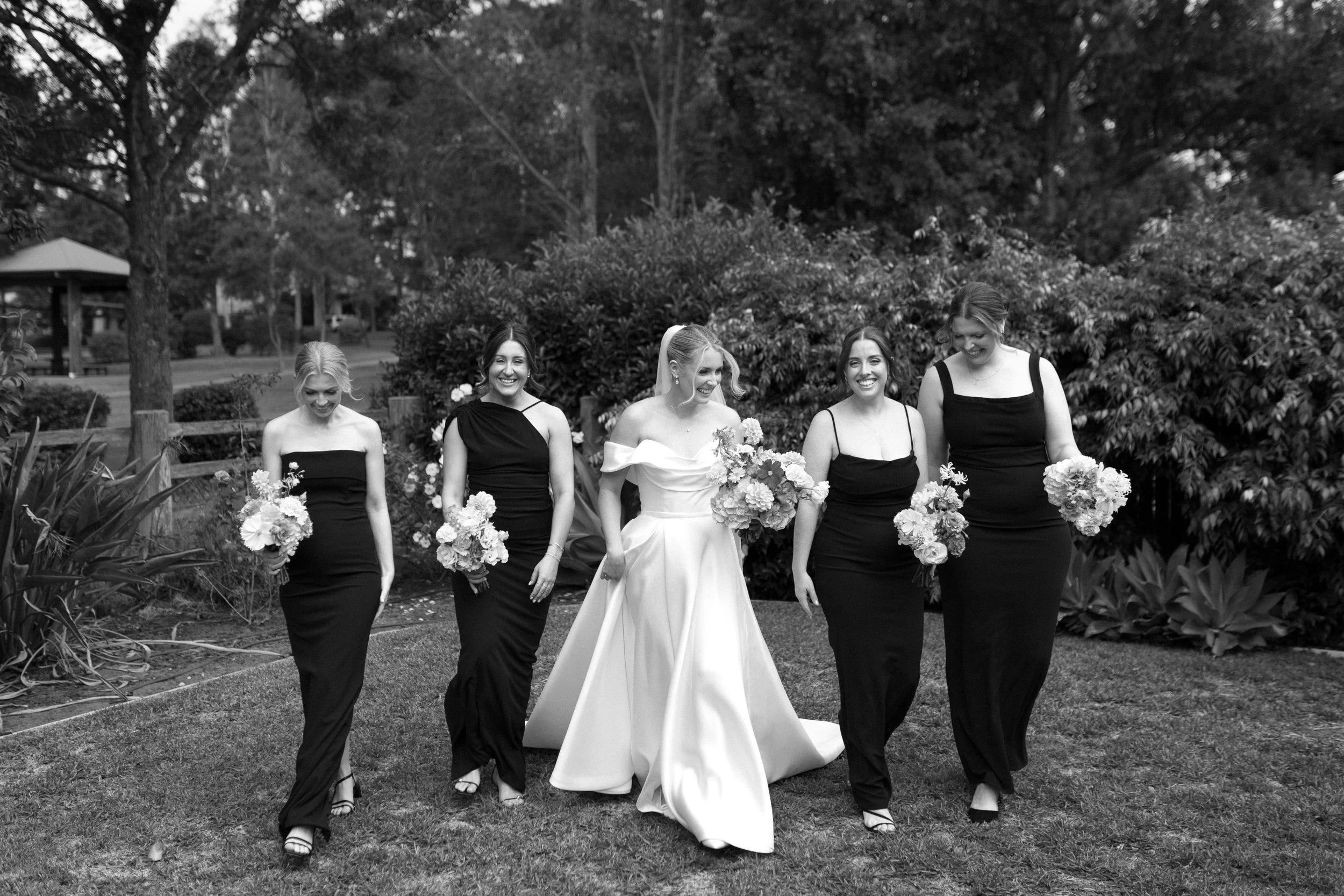 A bride and four bridesmaids walking outdoors in a garden, holding bouquets of flowers, with trees and bushes in the background, all smiling.