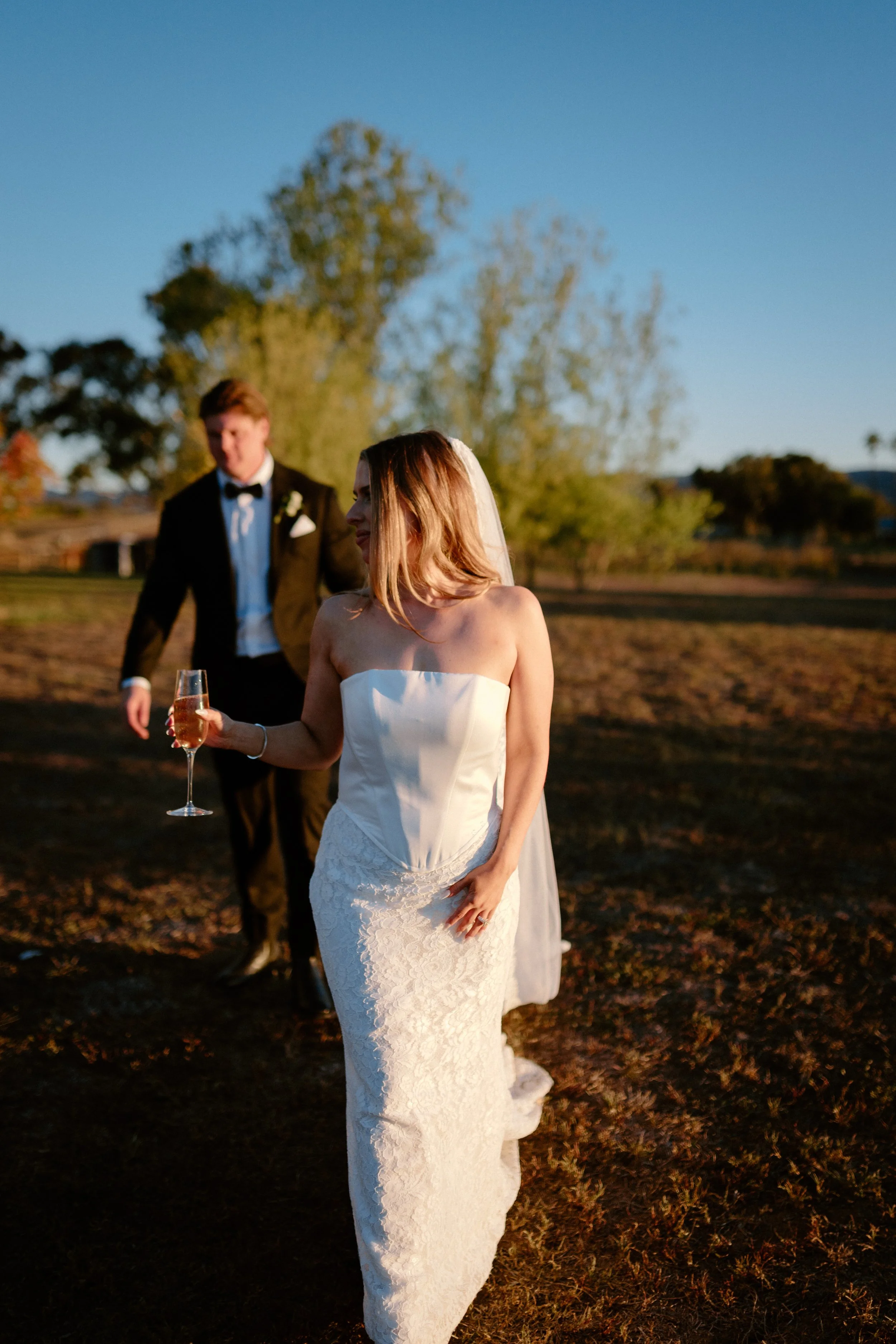 Bride in a white lace wedding dress holding a champagne glass standing outdoors during sunset, with groom in a black tuxedo in the background.