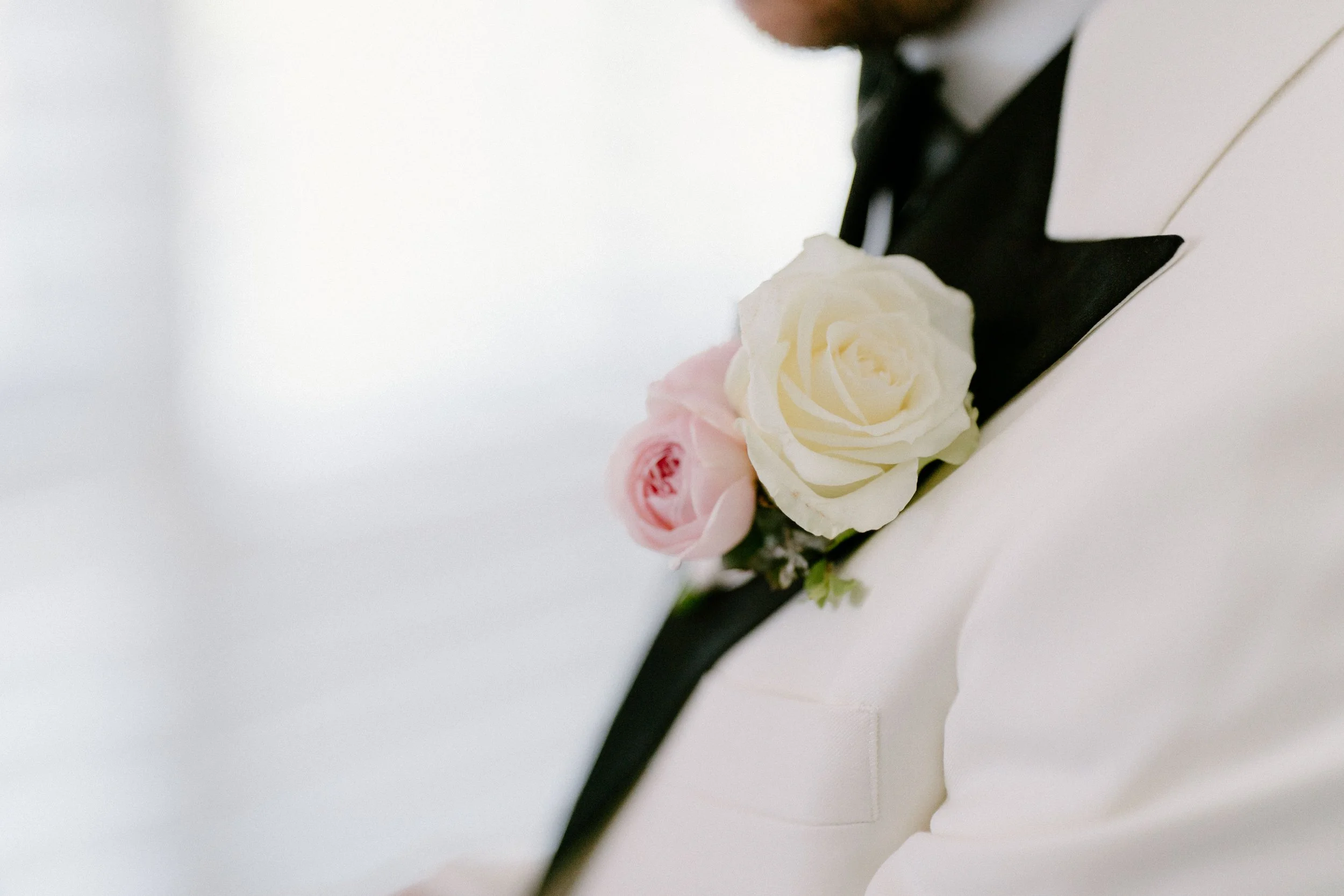 Close-up of a person dressed in formal attire with a white jacket and a black collar, wearing a boutonniere made of white and pink roses.