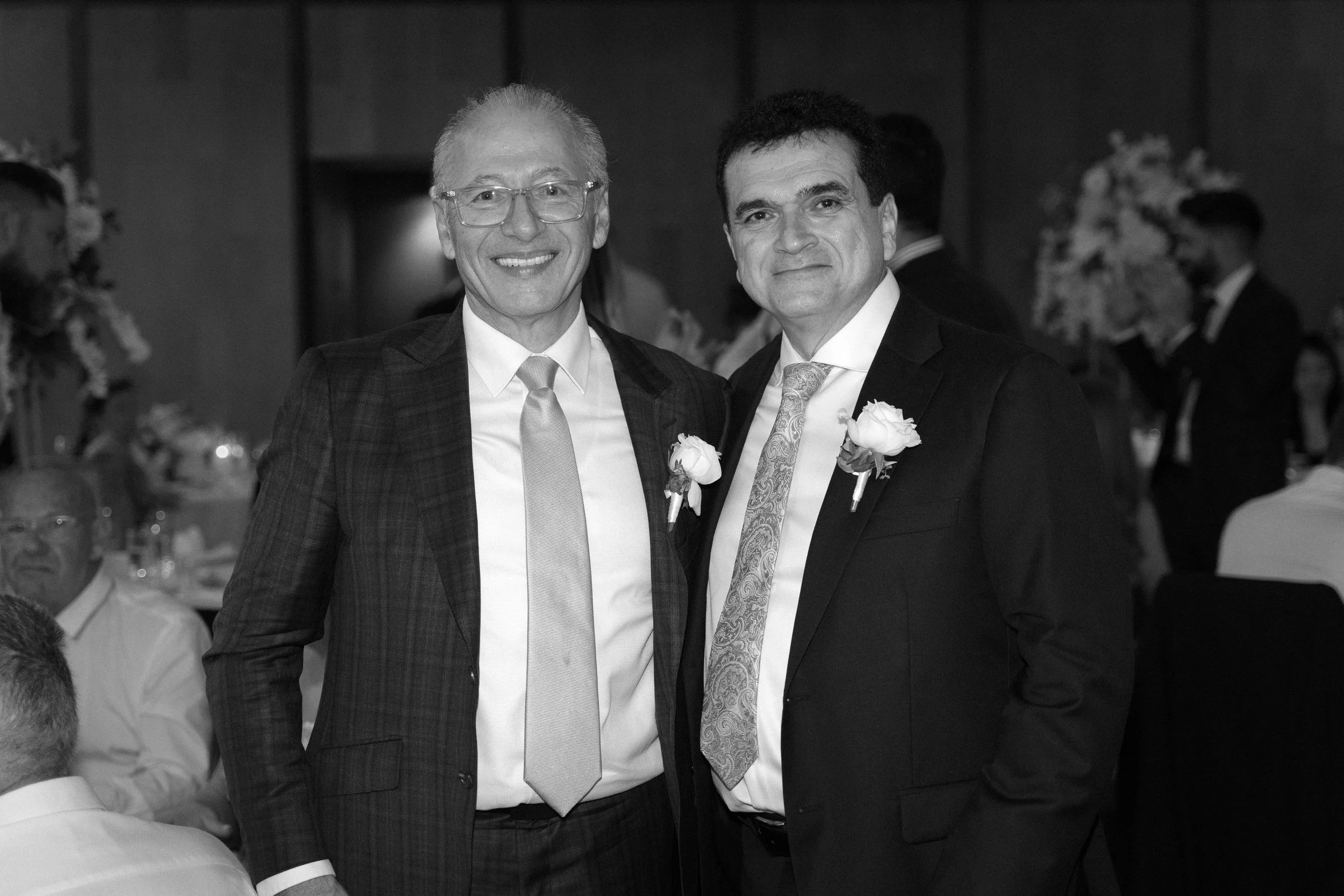 Two men in suits with floral boutonnières, smiling at a formal event, standing close together in a decorated banquet hall.