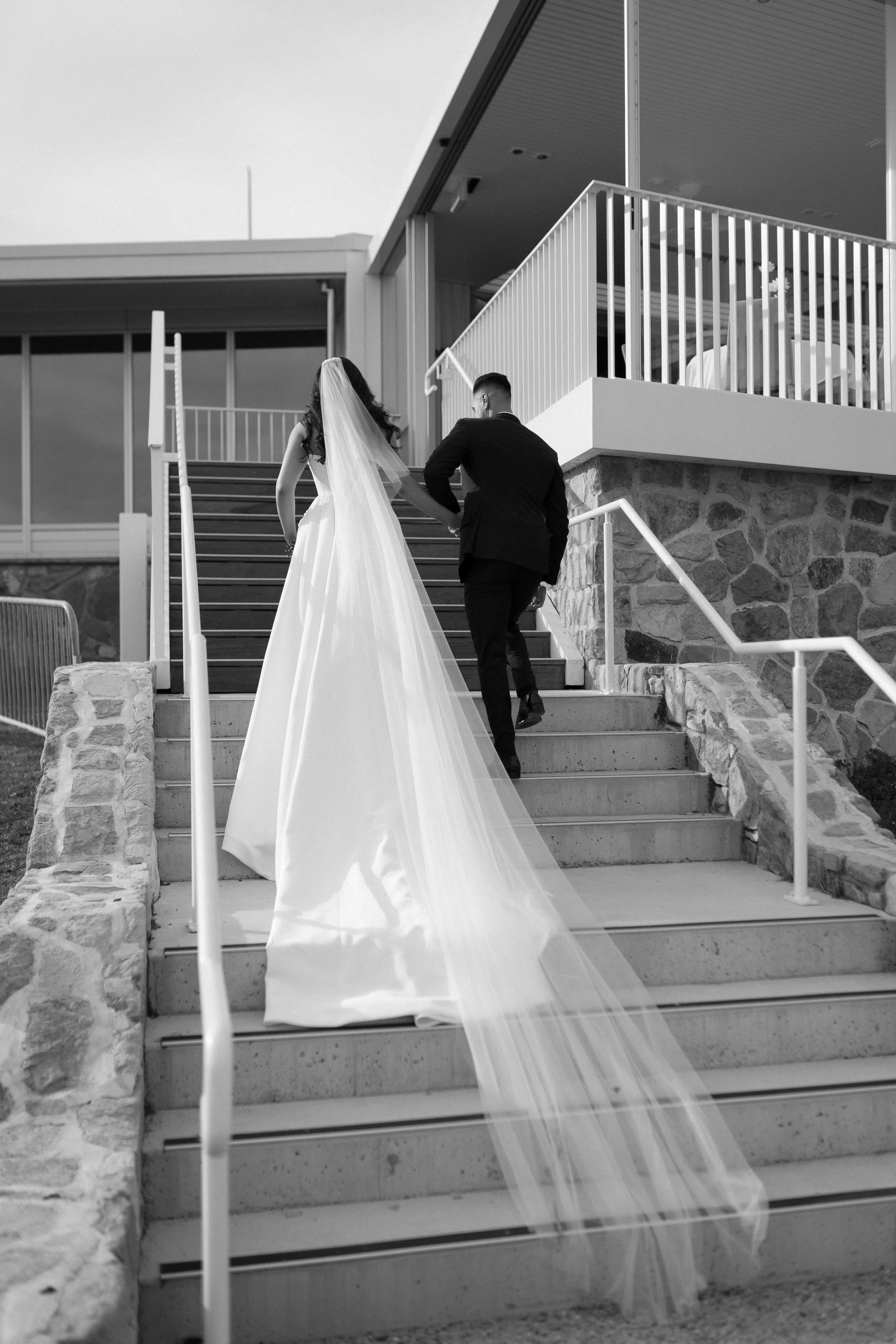 A bride in a wedding dress with a long veil and a groom in a suit ascending outdoor stairs towards a modern building.